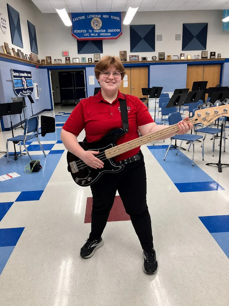 A girl with short brown hair and glasses, wearing a red shirt and black pants, standing in a school music room, smiling and holding a black electric bass guitar.