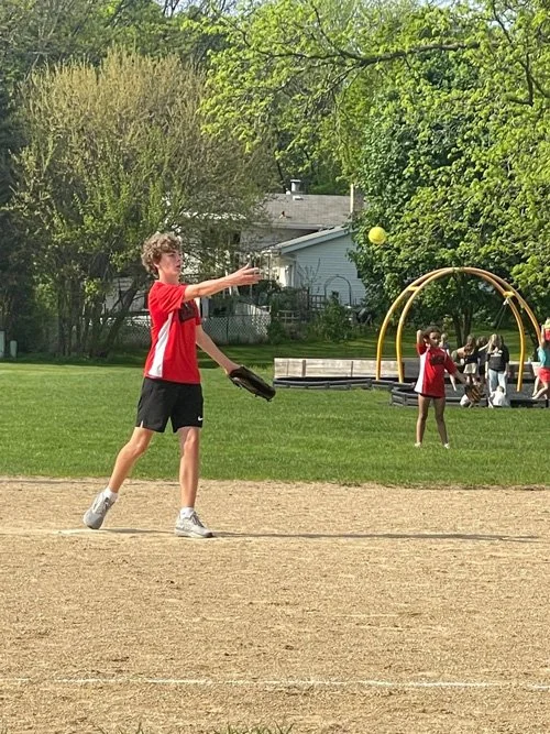A young boy playing baseball on a field with a glove, throwing a ball towards a girl in a red shirt who is standing in the grass, with playground equipment and trees in the background.