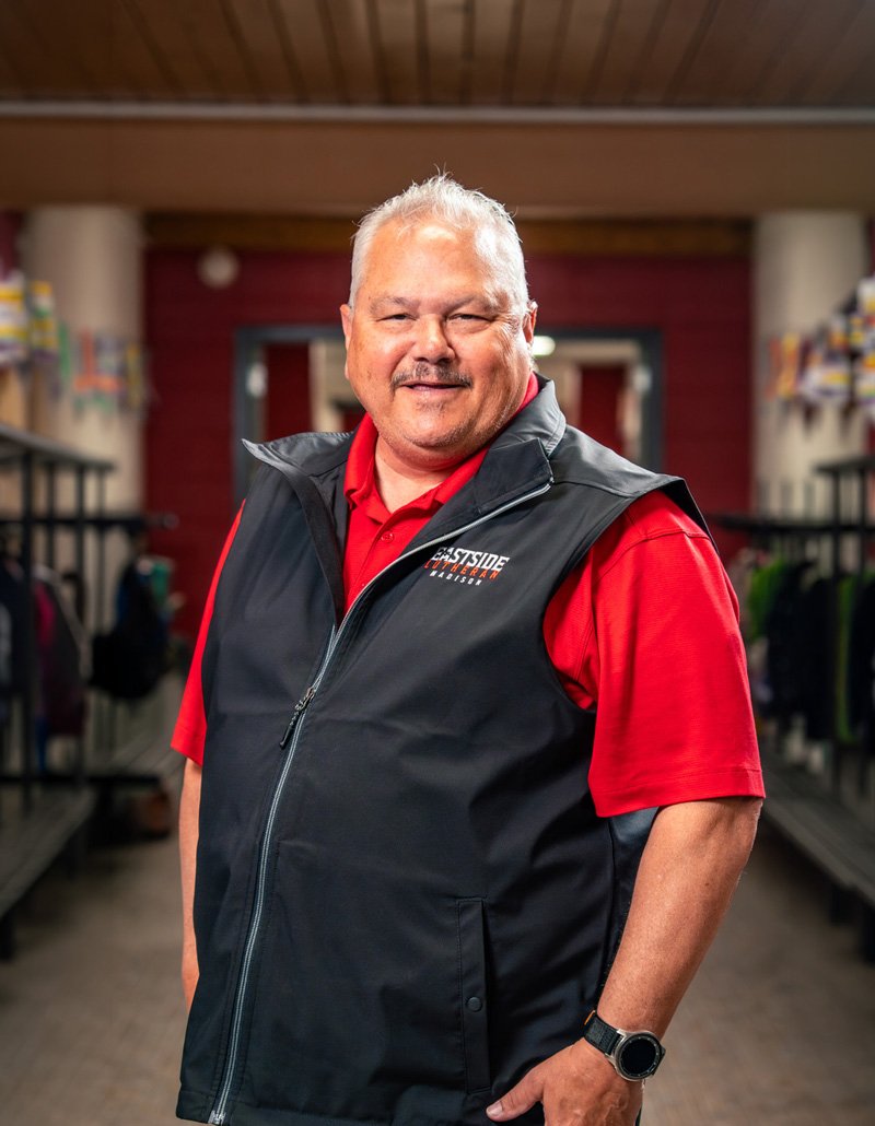 A middle-aged man smiling, wearing a red shirt and a black vest with a logo, standing in a store or warehouse aisle with lockers or storage units on the sides.