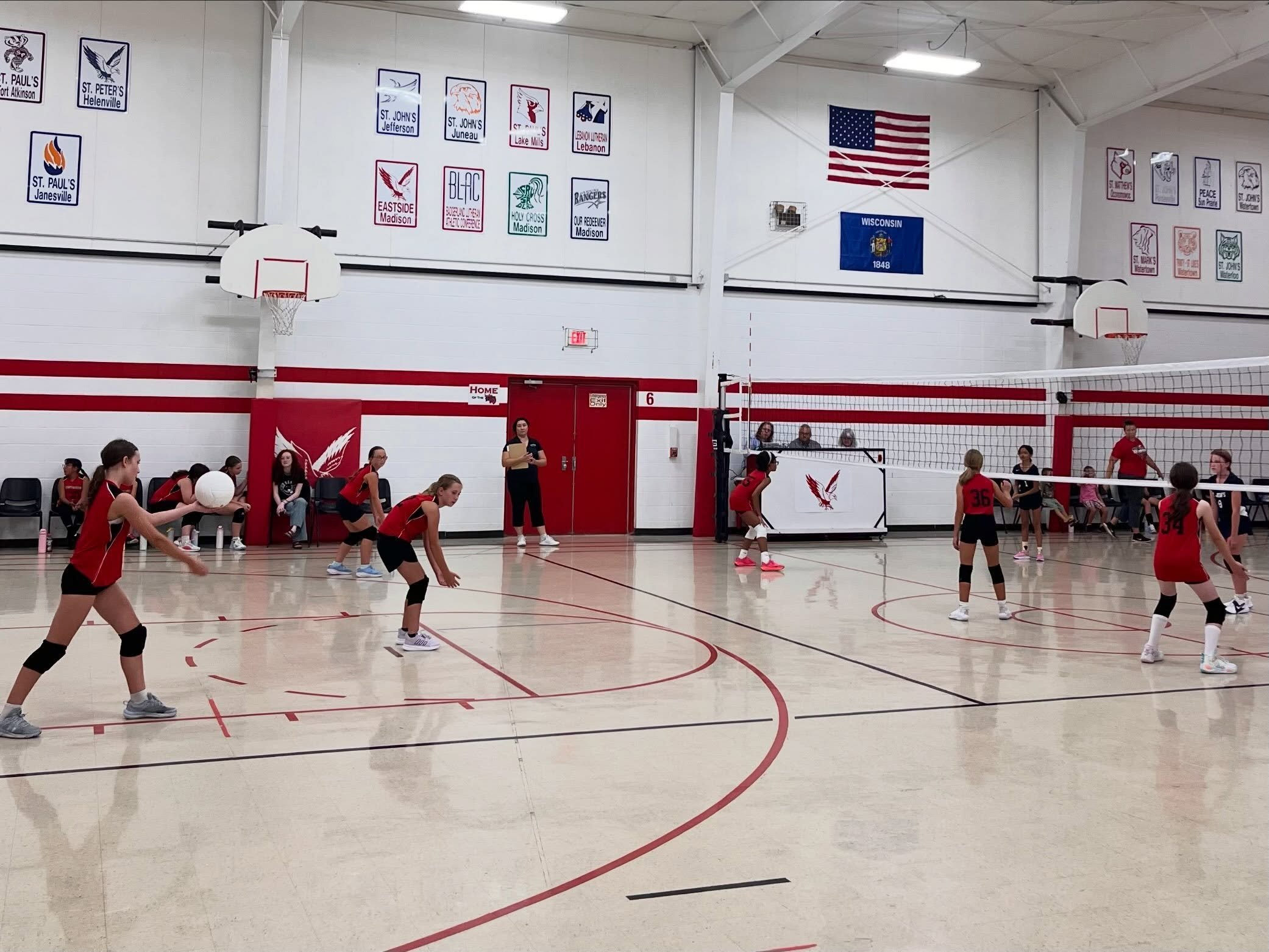 Indoor volleyball game with players in red uniforms on the court, a referee, and spectators seated on the sidelines. The gym has banners on the wall, including an American flag and a Wisconsin state flag.