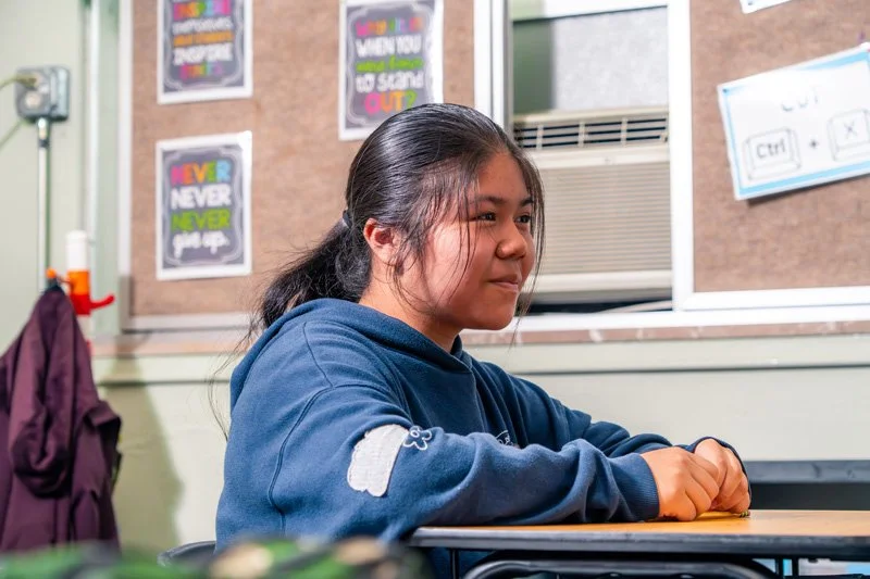 A young girl sitting at a desk in a classroom, wearing a blue hoodie, with classroom posters and a window in the background.