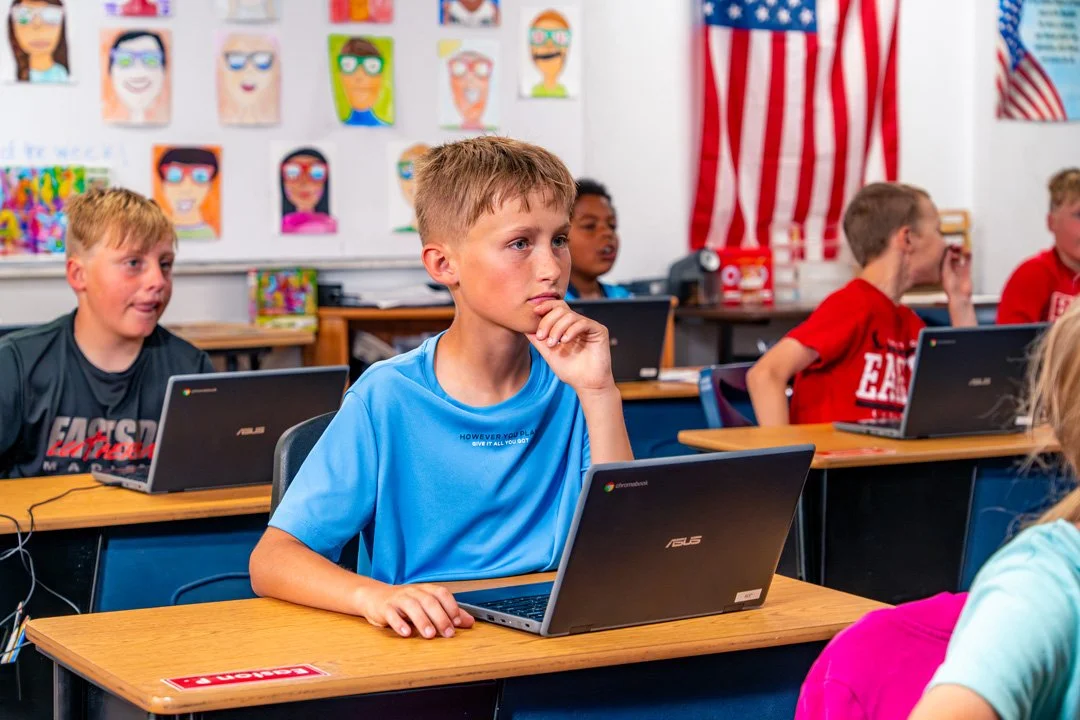 Young students sitting at desks in a classroom working on laptops, with colorful artwork and an American flag on the wall.