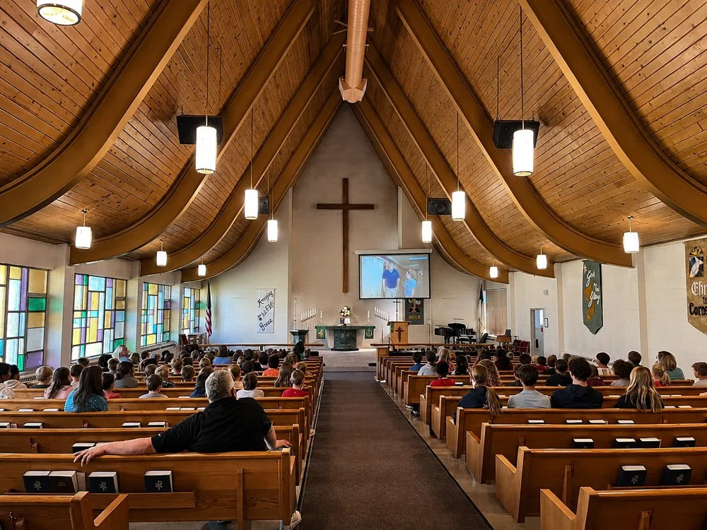 Inside a church with a congregation seated on wooden pews, stained glass windows on the left, a cross on the front wall, and a large screen displaying church service, with wooden ceiling beams and hanging lights.