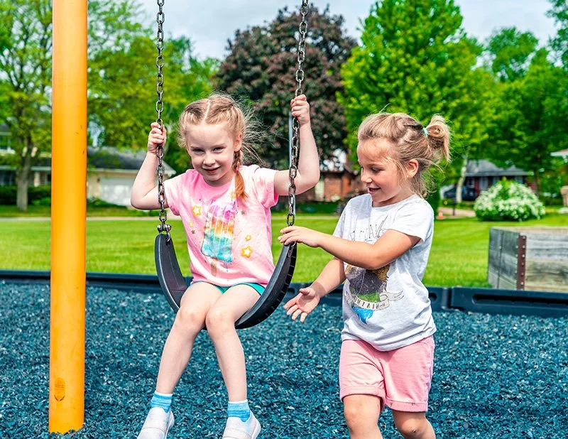 Two young girls playing on a swing set in a park on a sunny day, one girl sitting on the swing and the other standing beside her.