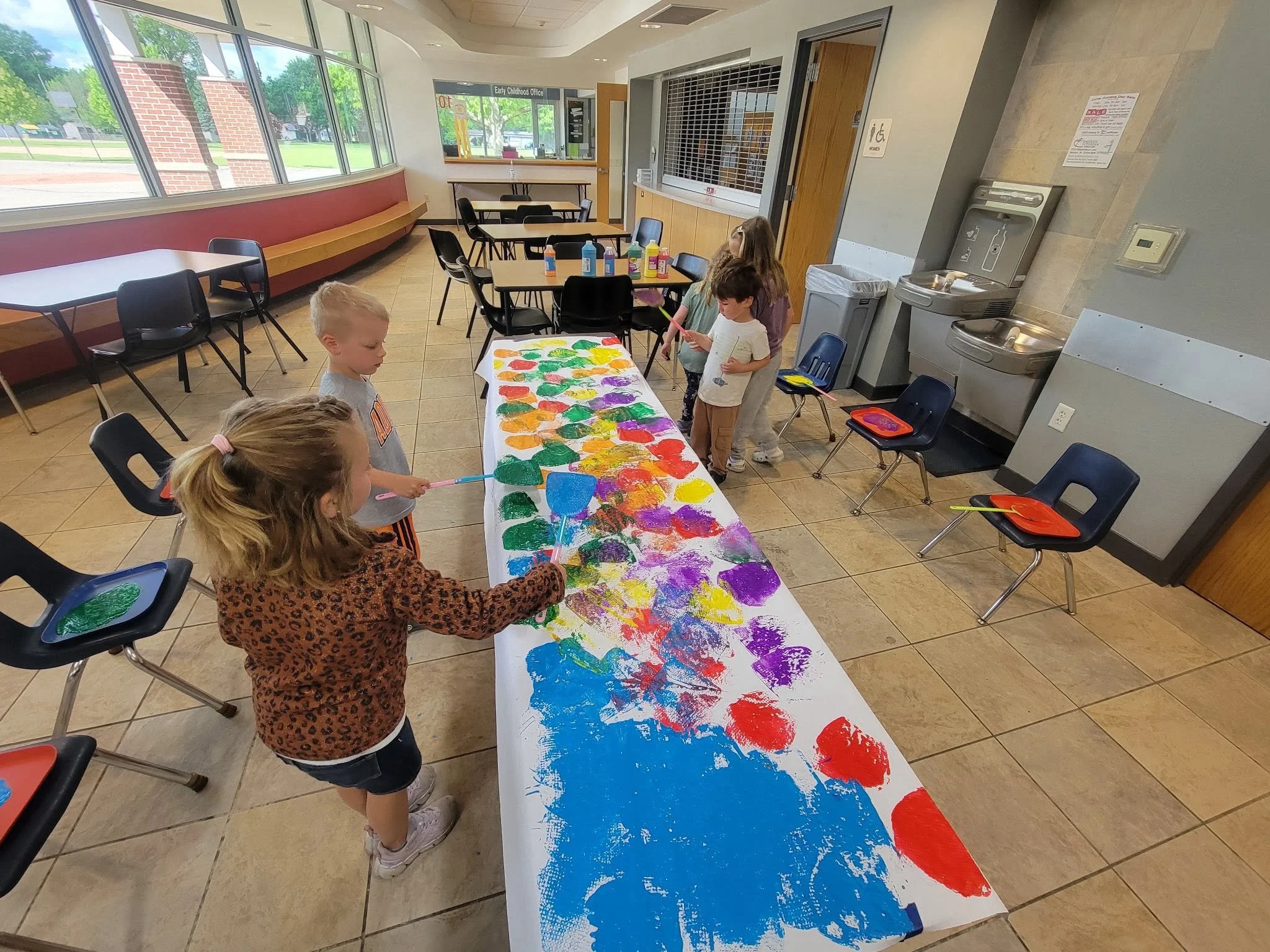 Children painting on a long strip of paper during an indoor art activity, with bottles of paint and paintbrushes visible, in a room with large windows and a water fountain.