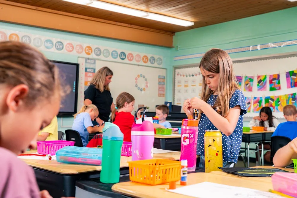 A classroom with young children organizing school supplies, some preparing to start a lesson, with posters and decorations on the walls.