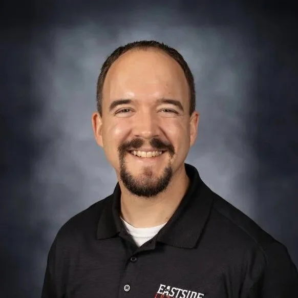 Headshot of a smiling man with short brown hair and a beard, wearing a black polo shirt with a logo on the chest, against a dark, gradient background.