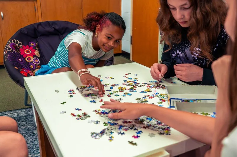 Children working on a puzzle at a table in a room with wooden cabinets and a carpeted floor.