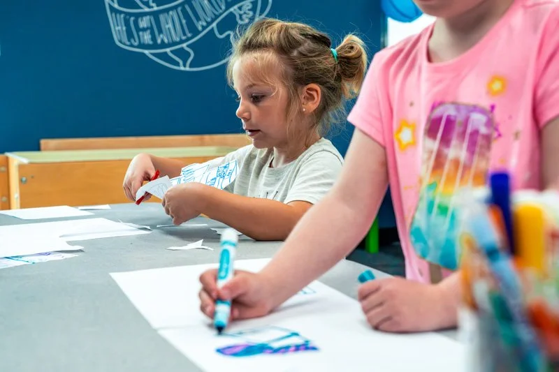 Two young girls engaged in drawing and coloring activities at a table in a classroom.