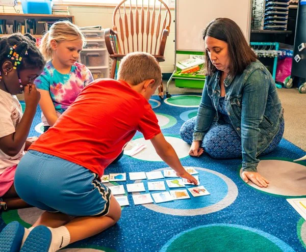 A woman with dark hair and a denim jacket sitting on a blue patterned rug, with three children around her, playing an educational card game on the rug in a classroom.
