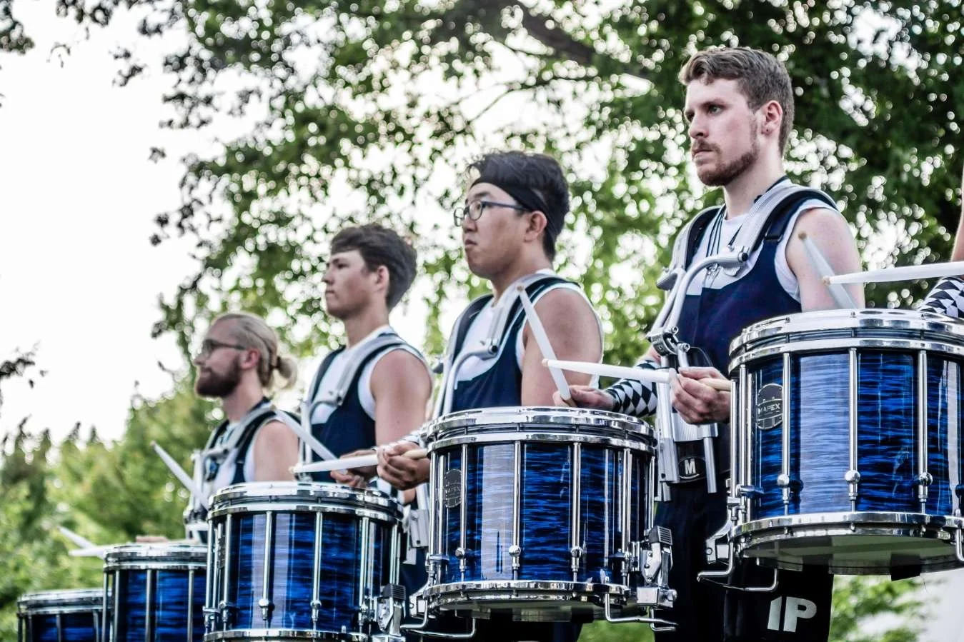 A group of five male drummers dressed in athletic attire standing outdoors with trees in the background, holding drumsticks and positioned behind blue drums.