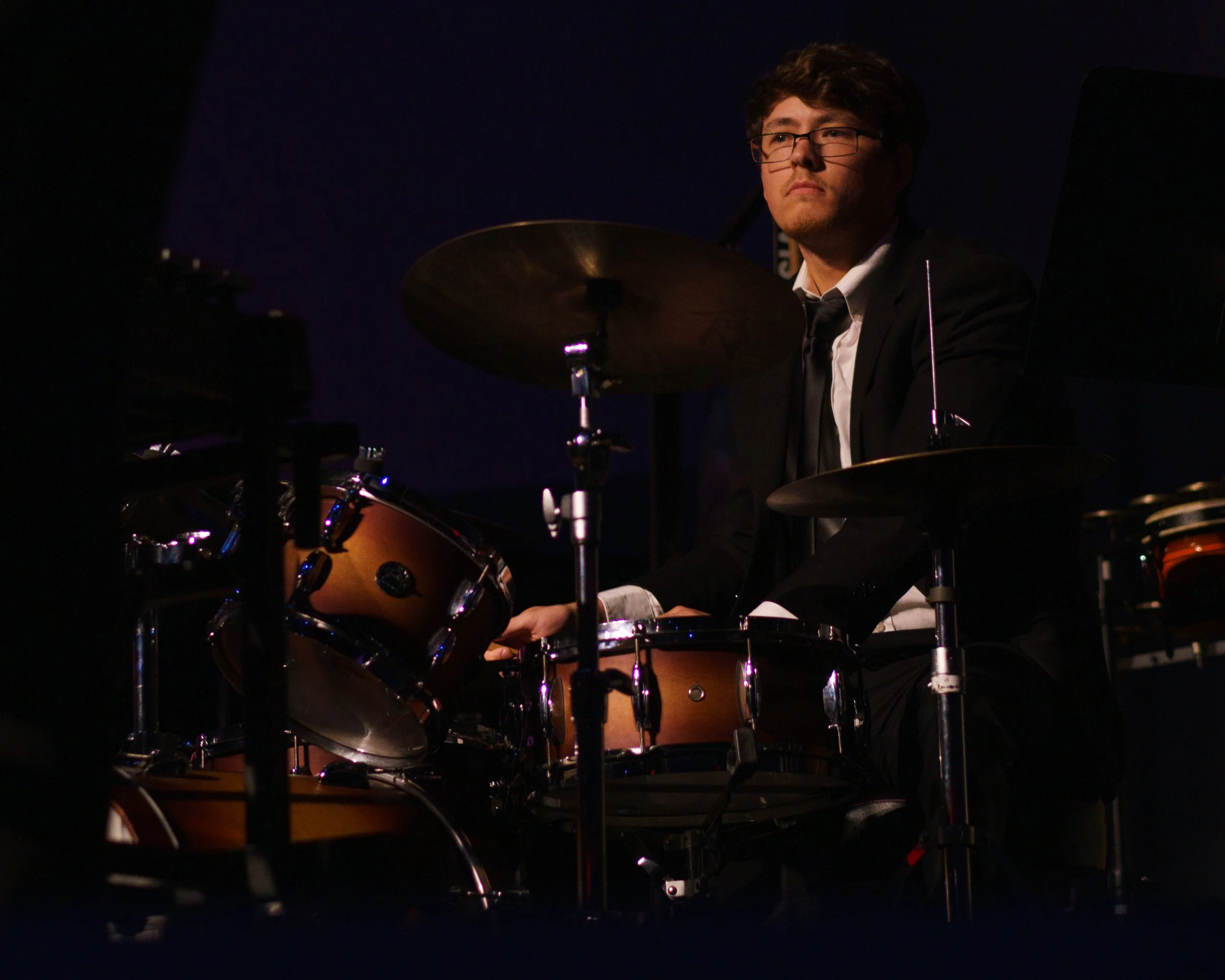 A man with glasses playing drums on a dark stage.