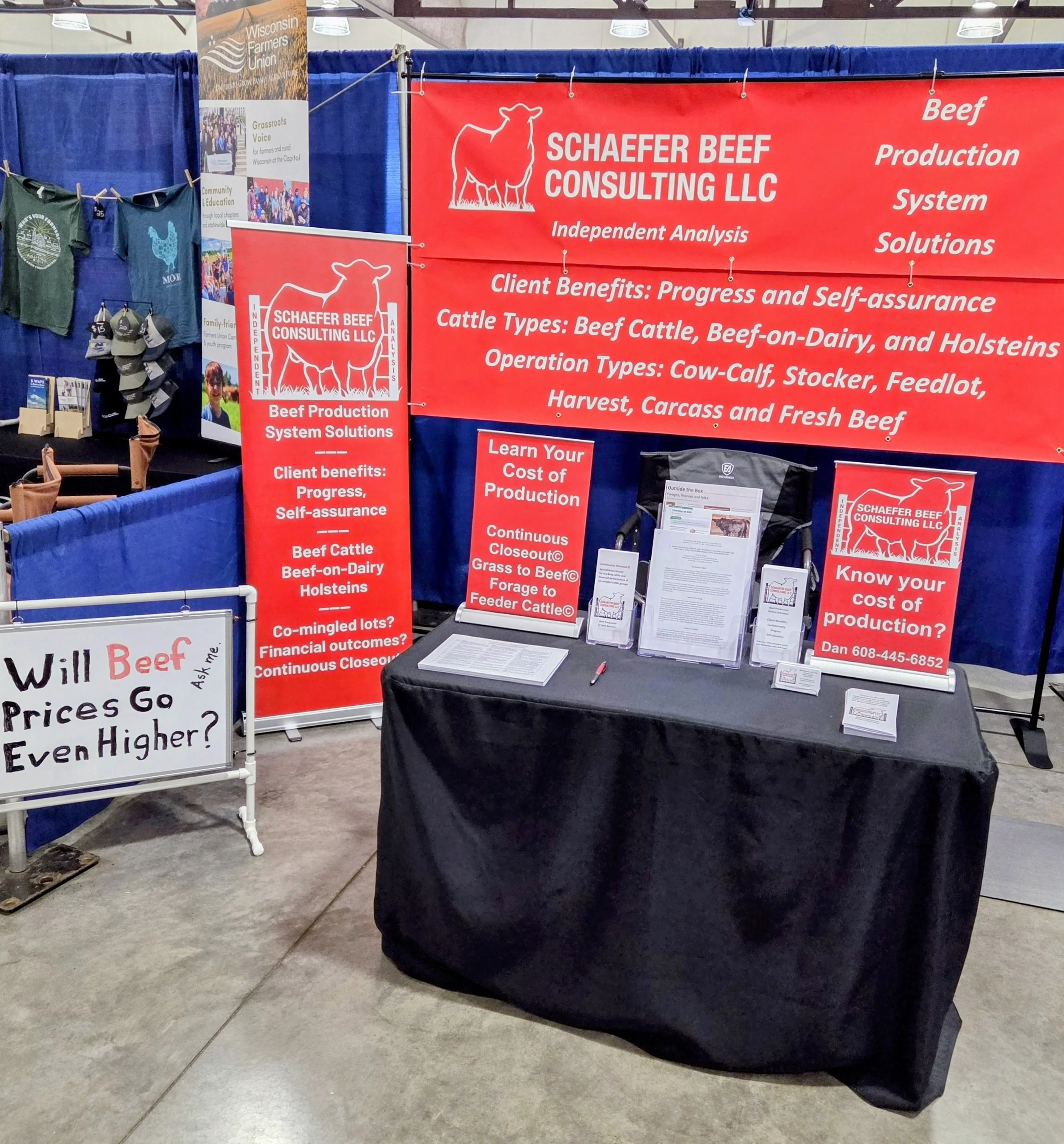 Booth for Schaefer Beef Consulting LLC at a convention, with red banners displaying information about beef production system solutions, and client benefits, and a table with pamphlets and business cards.