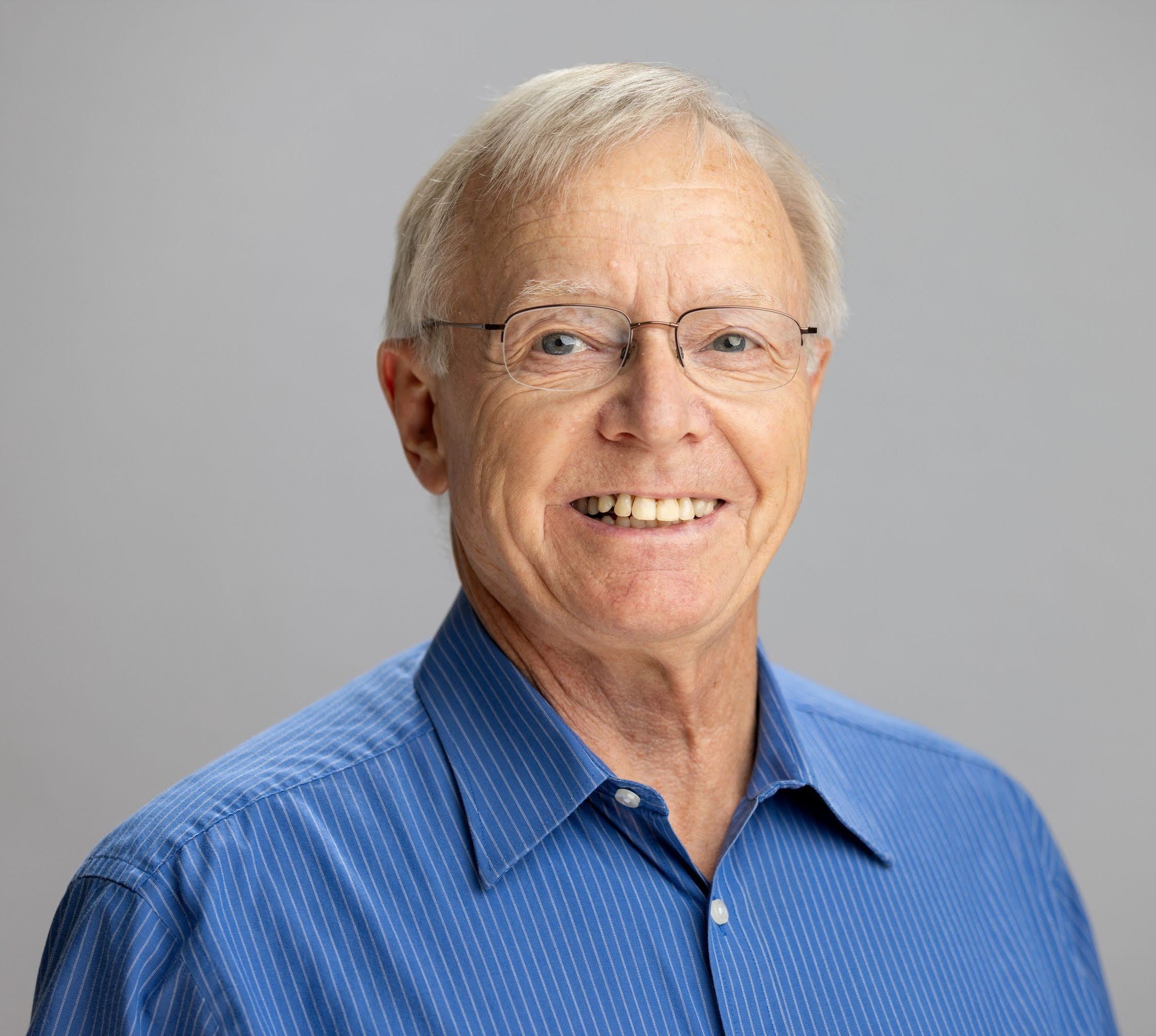 Close-up of Dan Schaefer with glasses, short gray hair, smiling and wearing a blue pinstripe shirt.
