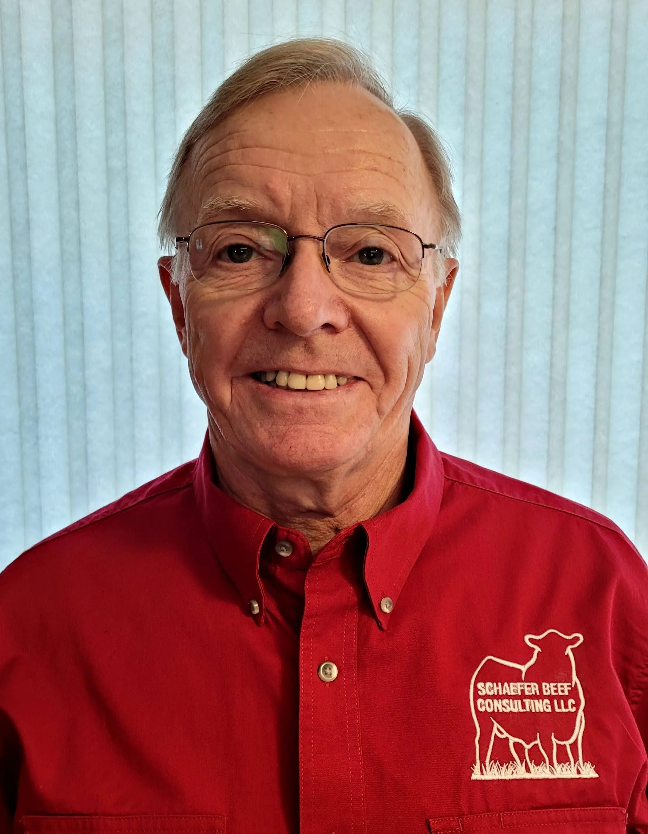 A man wearing glasses and a red button-up shirt with a logo reading 'Schafer Beef Consulting LLC' and a graphic of a beef bull, standing in a grass pasture.