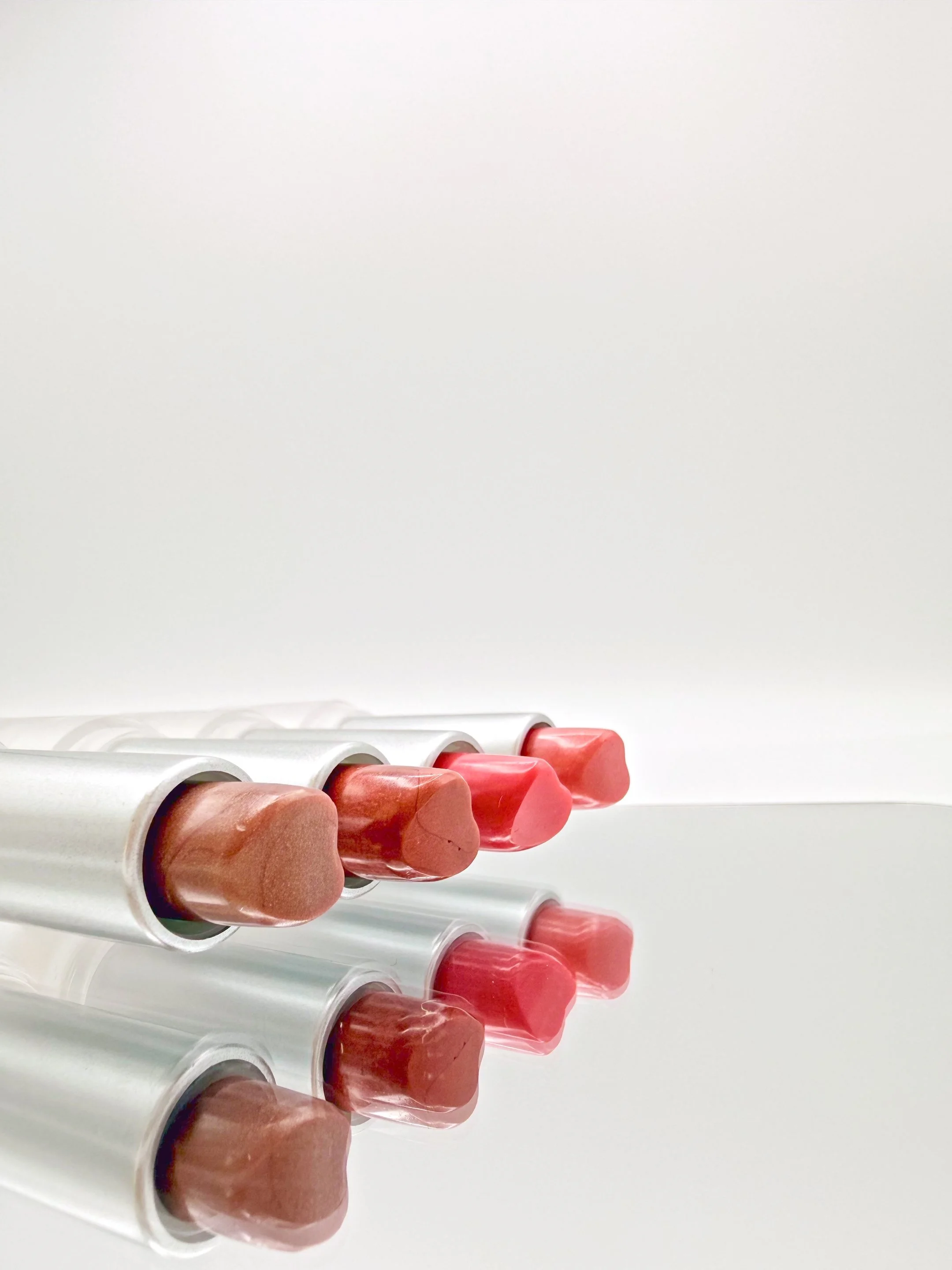 A row of heart-shaped lipsticks/lipbalms with beige and pink shades arranged in transparent tubes on a white surface.