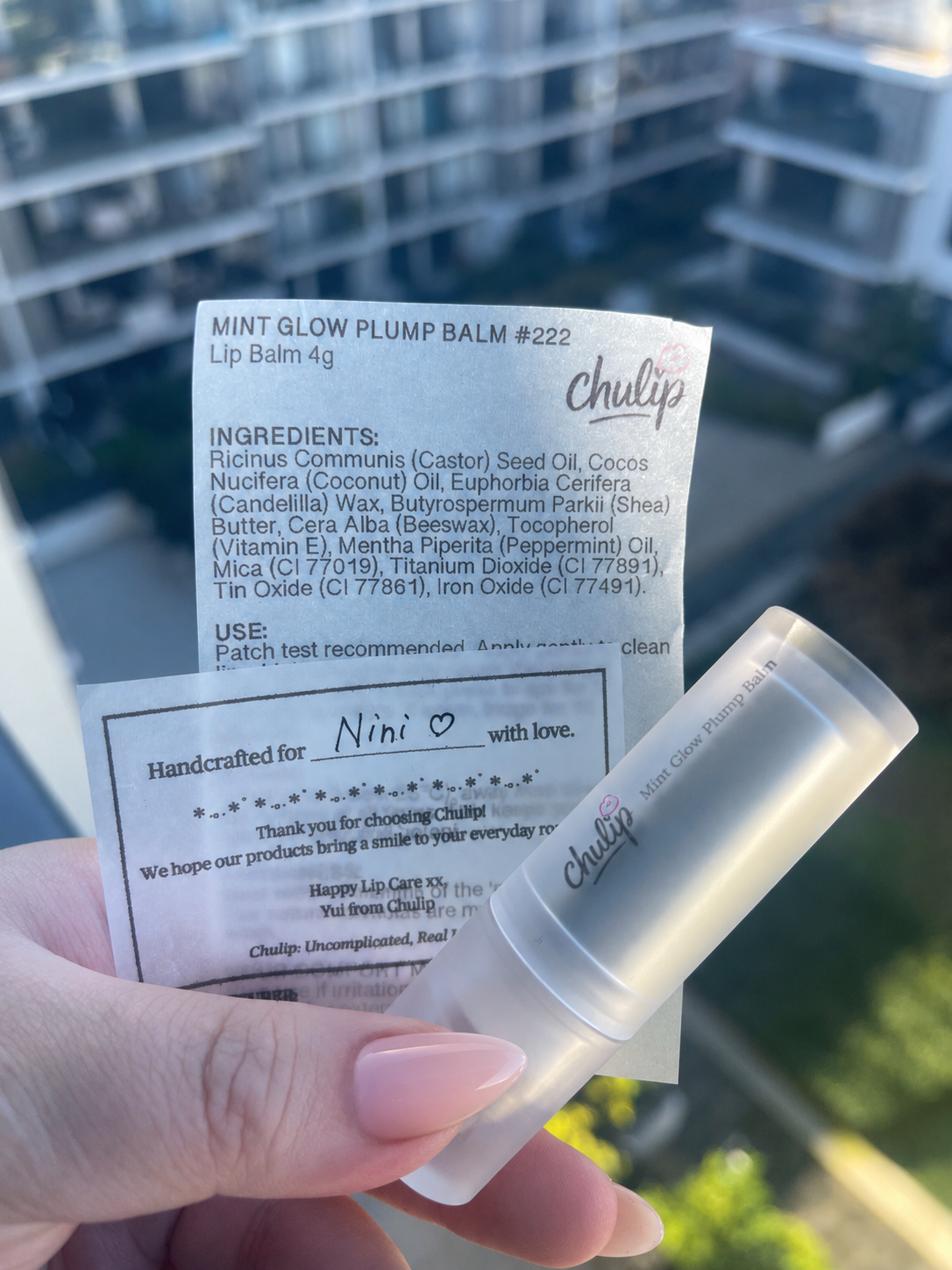 A hand holding a Mint Glow Balm and a product packaging label with ingredients, instructions, and a note to Nini, against a background of a building with balconies.