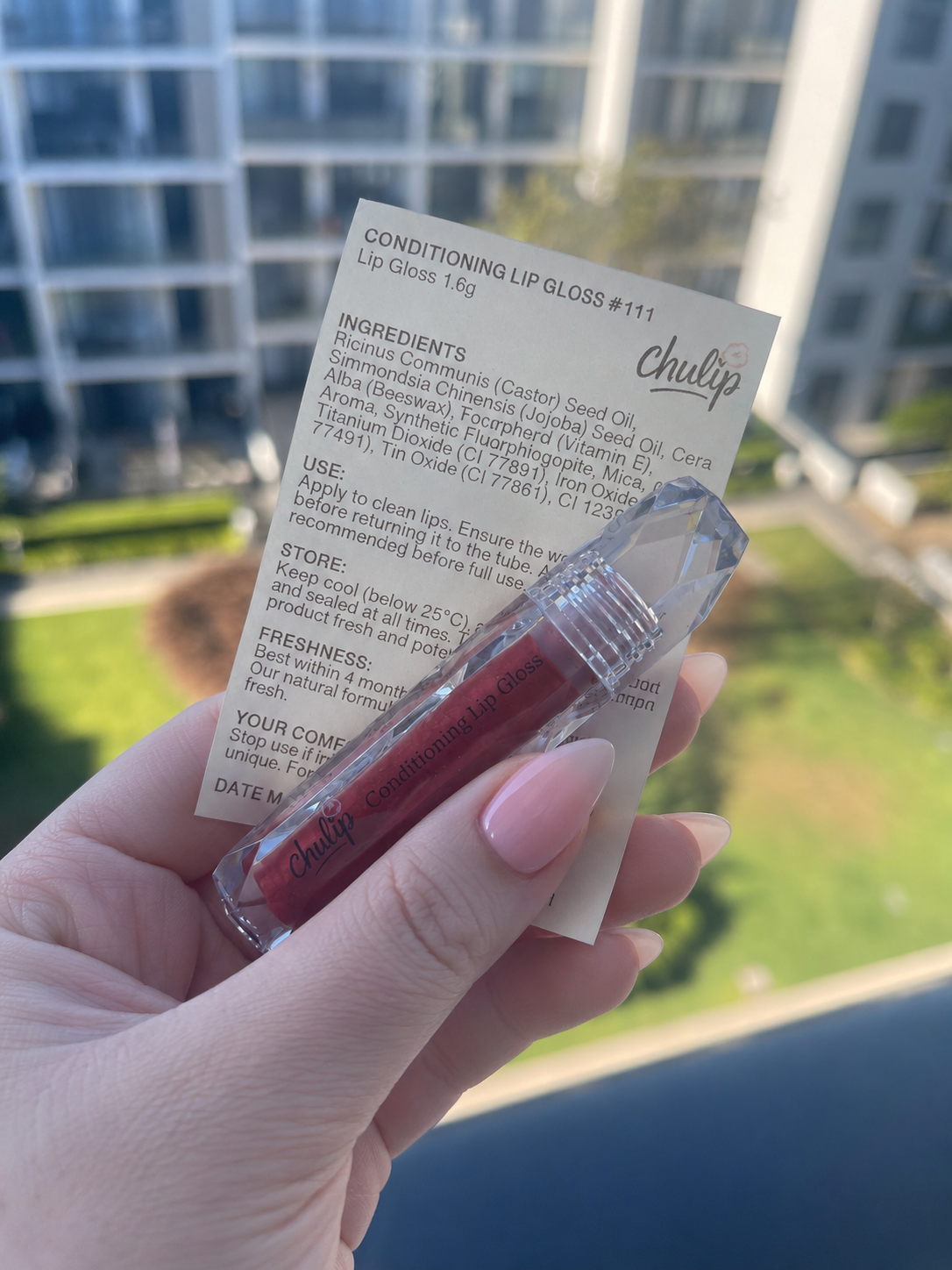 A person holding a small container of conditioning lip gloss and a label with ingredients and usage instructions, with a blurred cityscape background.
