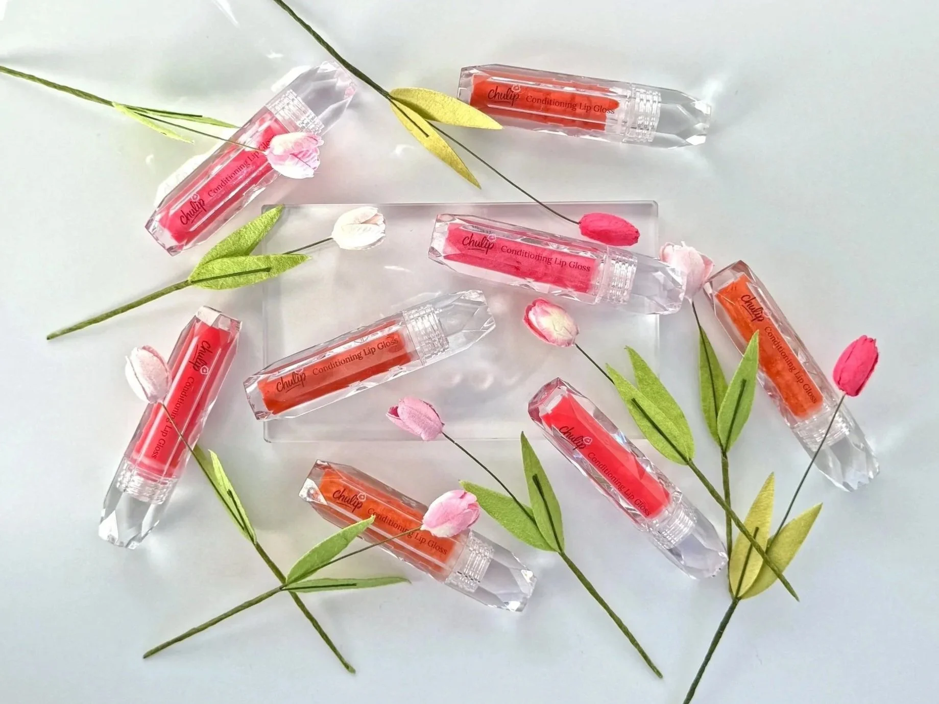 Multiple tubes of CHULIP Conditioning Lip Gloss surrounded by pink and white artificial tulip flowers and green leaves on a white background.