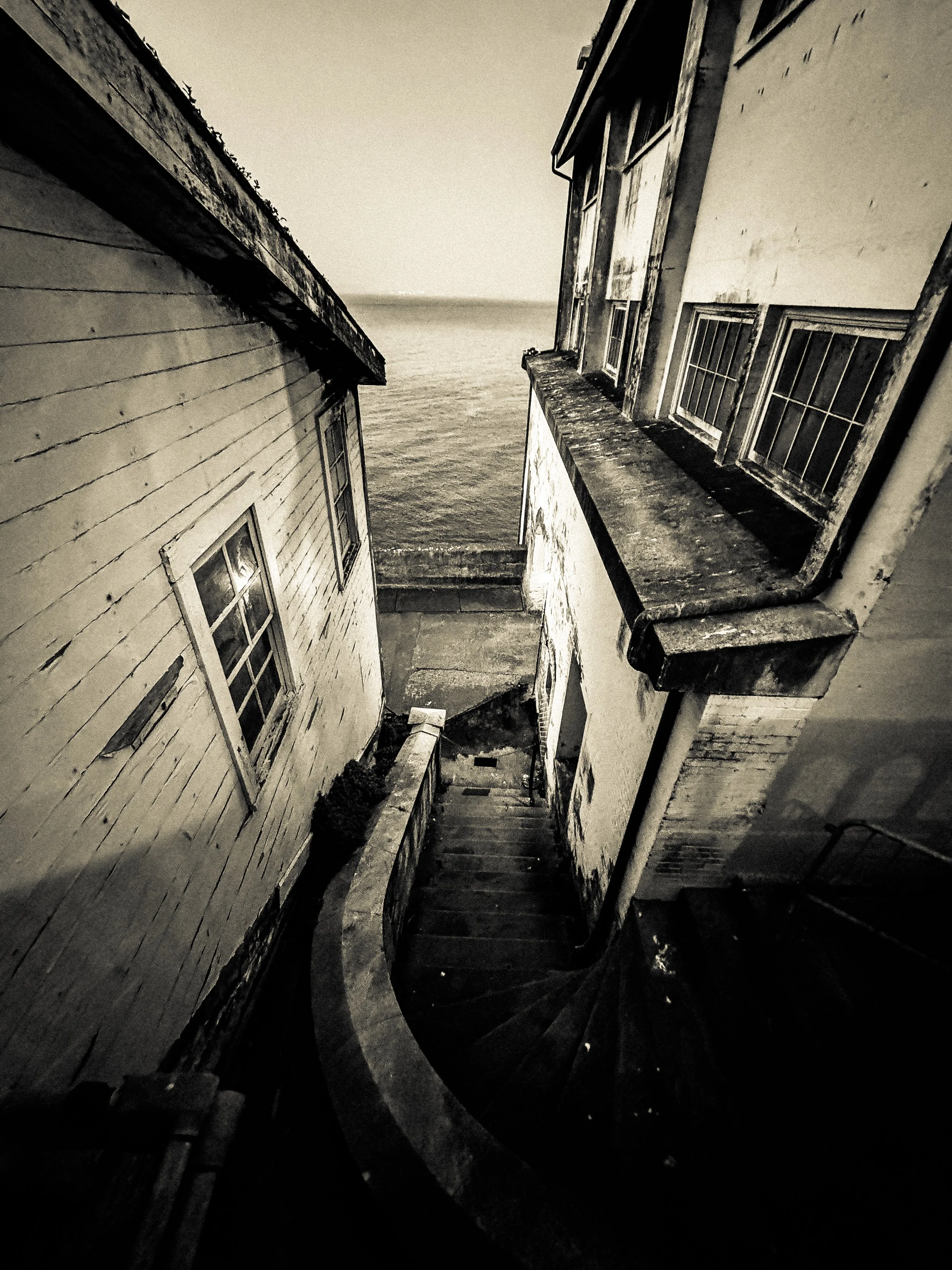 View of two weathered, wooden houses with windows on a narrow alleyway, leading to the ocean in the background, in black and white