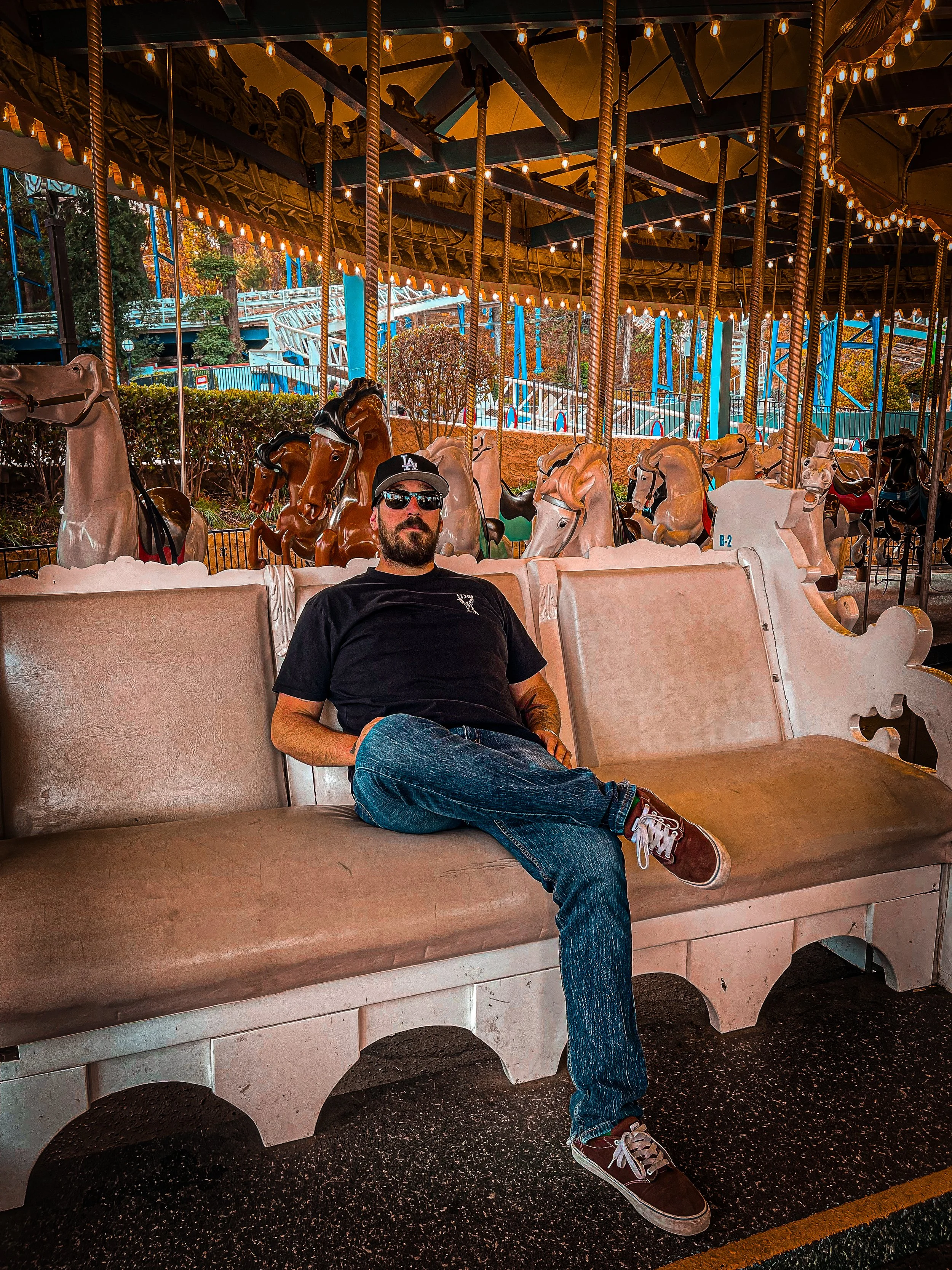 A man with a beard wearing sunglasses, a black t-shirt, blue jeans, and brown sneakers, sitting on a carousel bench at an amusement park, with a carousel horse behind him and a roller coaster in the background.