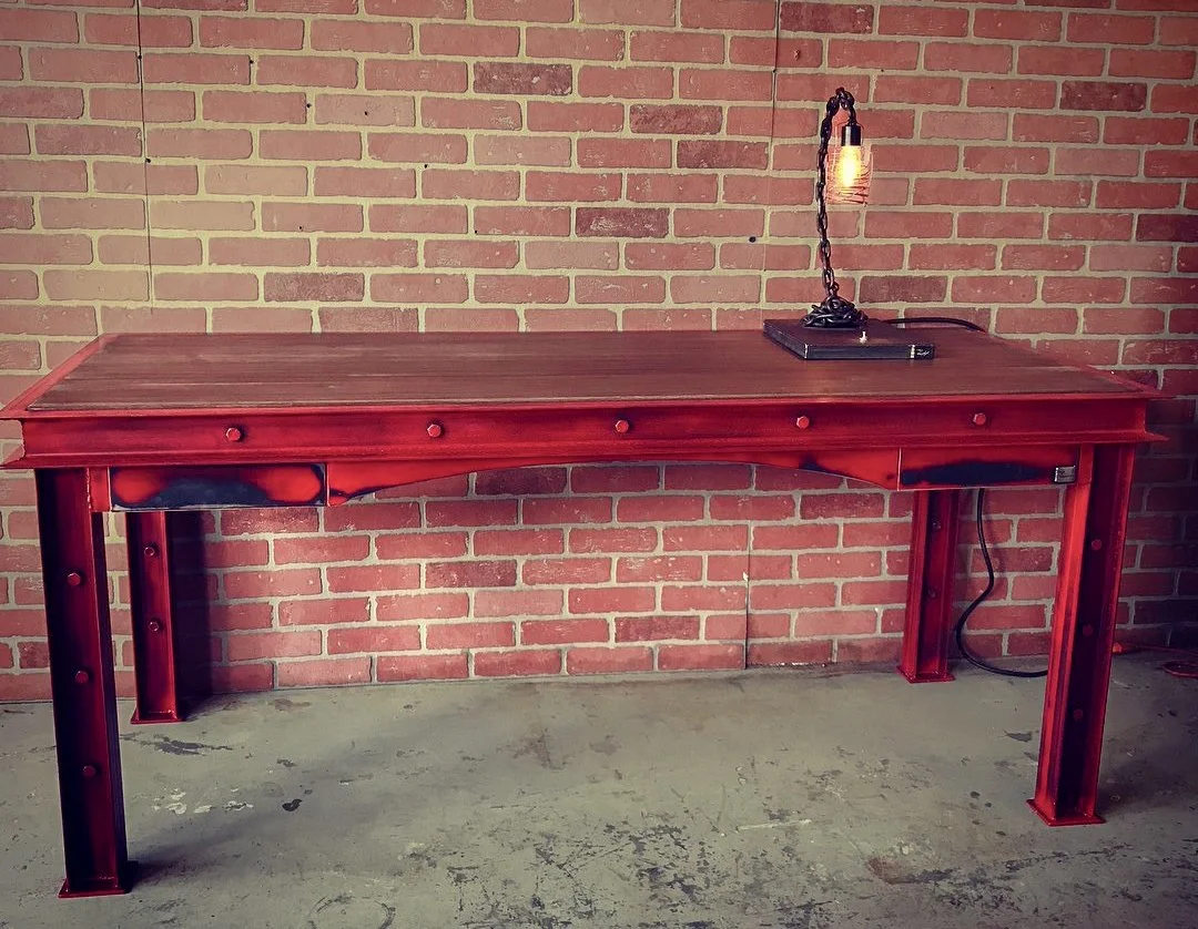 A large fabricated metal and wood industrial desk with a reddish finish sits against a brick wall. A single table lamp with an exposed Edison bulb and black chain is on the right side of the desk, plugged into an electrical outlet.