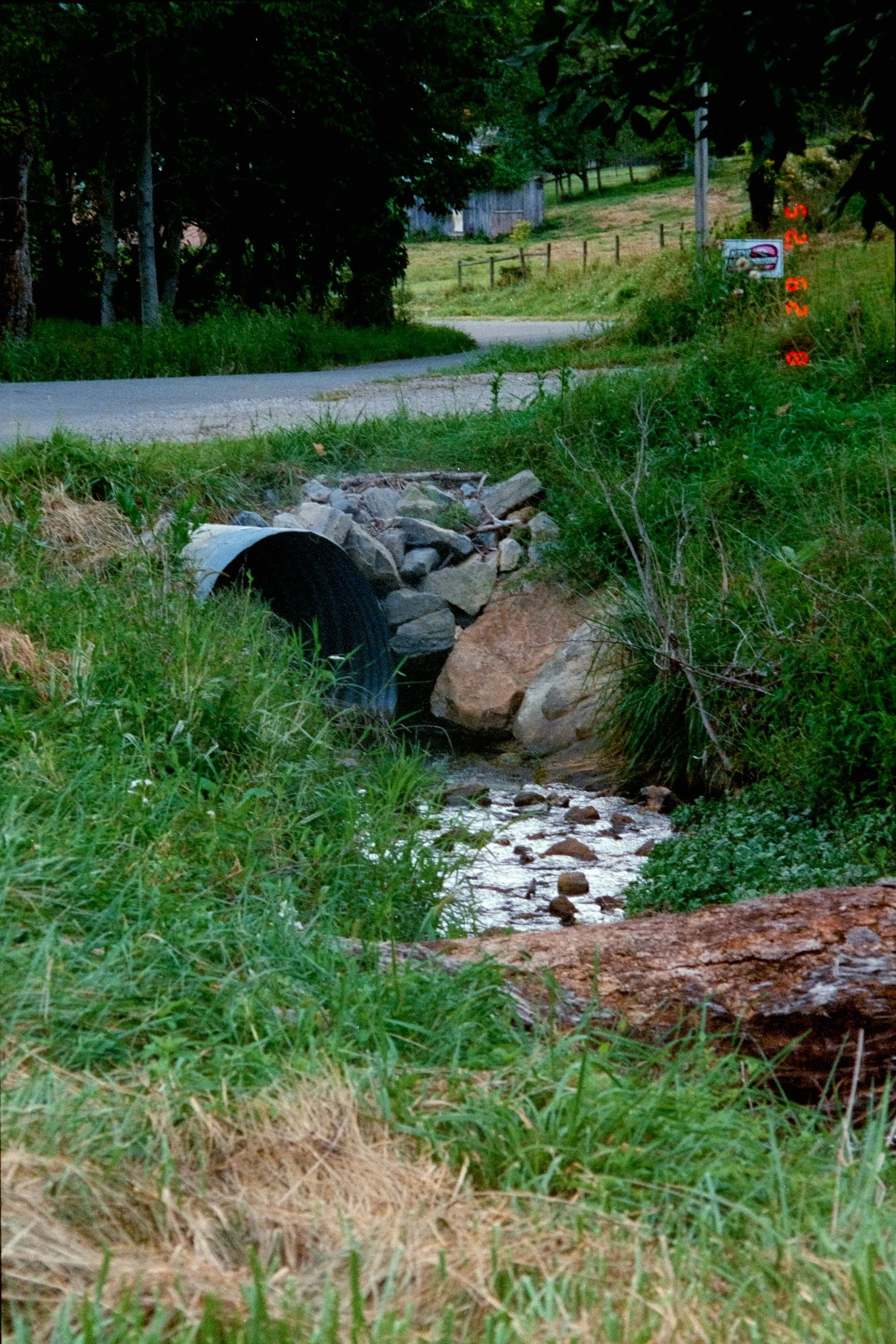 A small stream flowing through green grass and rocks in a rural area, with a pipe partially submerged in the stream, and trees and a wooden fence in the background.