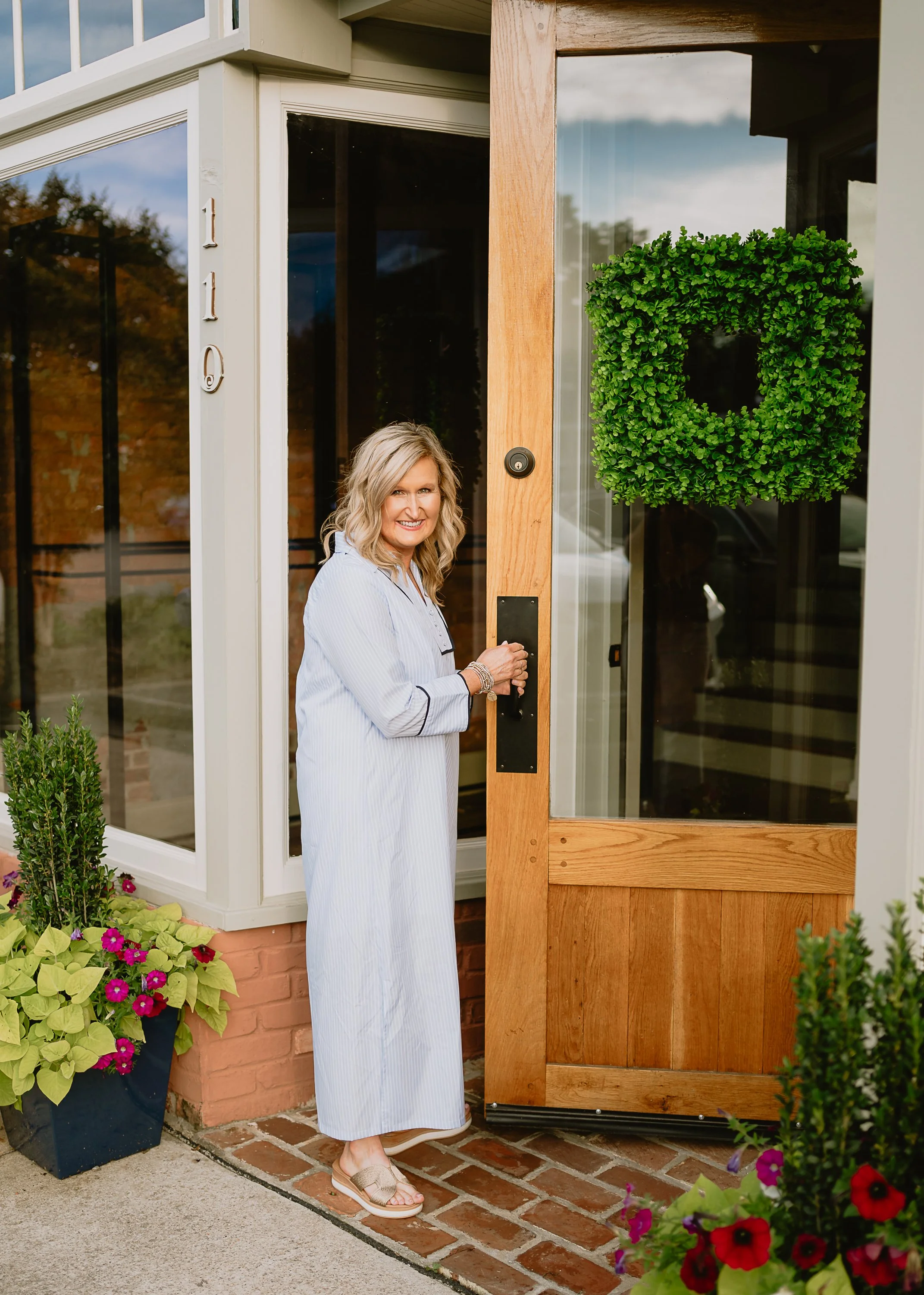 Woman in a striped nightgown standing at a wooden door with a green wreath, outside a house with potted flowers and plants.