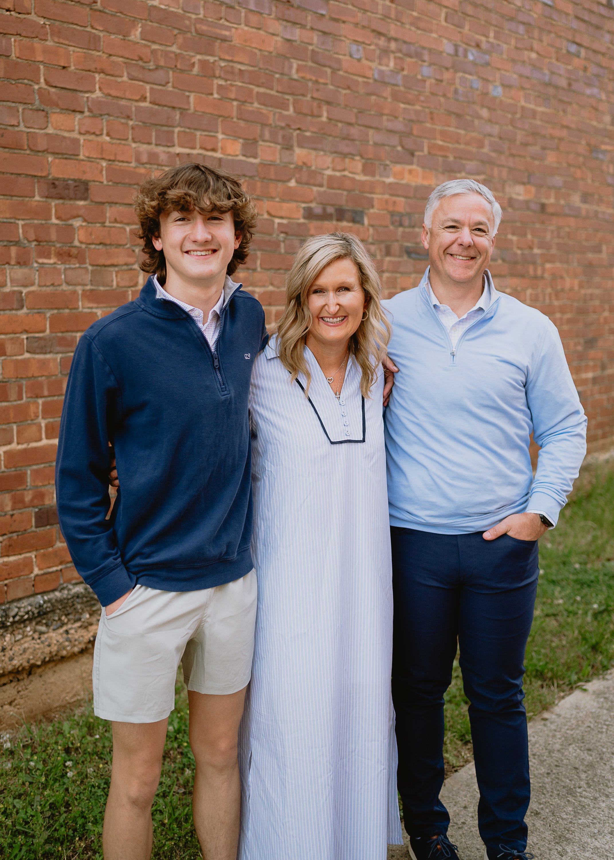 Family portrait of three people standing outdoors in front of a brick wall. The group includes a young man, a middle-aged woman, and a middle-aged man, all smiling and dressed casually.