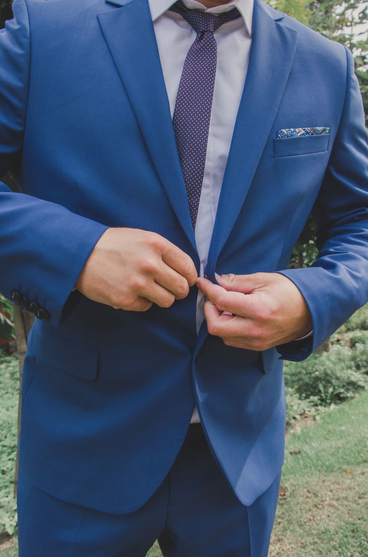 A man in a blue suit buttoning his jacket, with a white shirt, a patterned tie, and a pocket square in his breast pocket, outdoors.