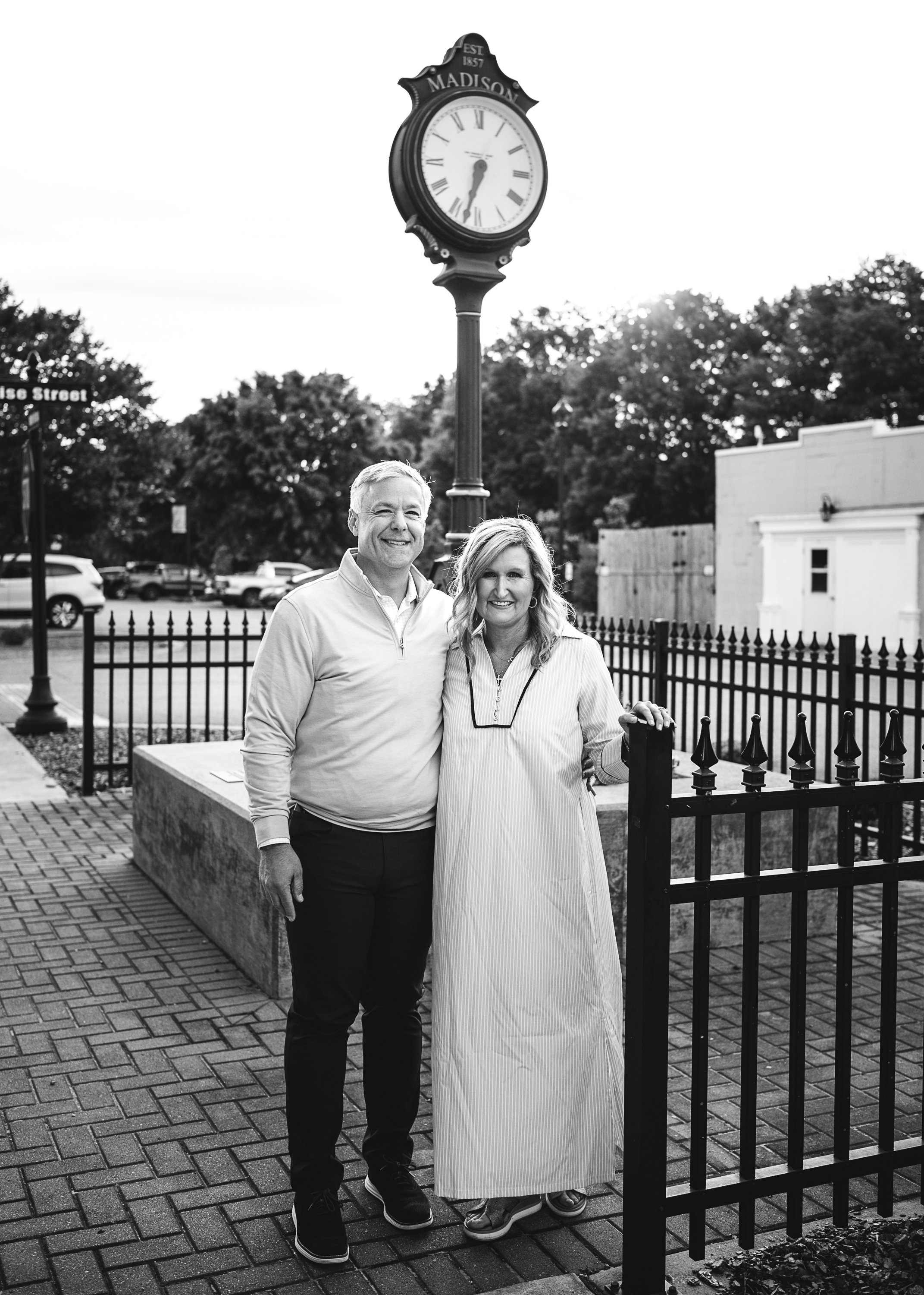 A smiling man and woman standing side by side outdoors near a black fence, with a street clock behind them showing approximately 6:20, trees, parked cars, and buildings in the background, in a black and white photo.