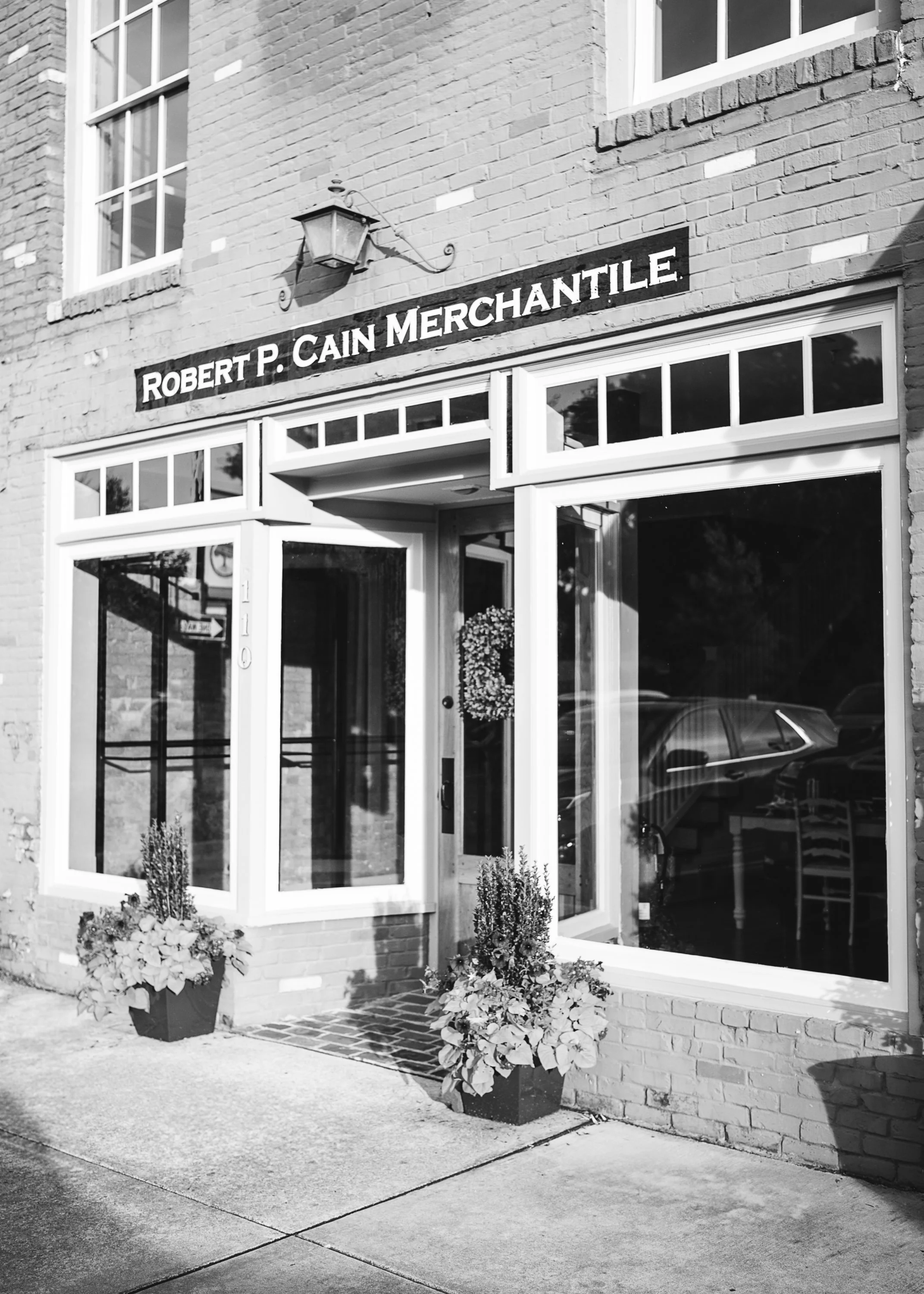 Storefront with a sign reading 'Robert P. Cain Merchantile' above large glass windows and a door. There are flower pots outside with plants, and a brick sidewalk.
