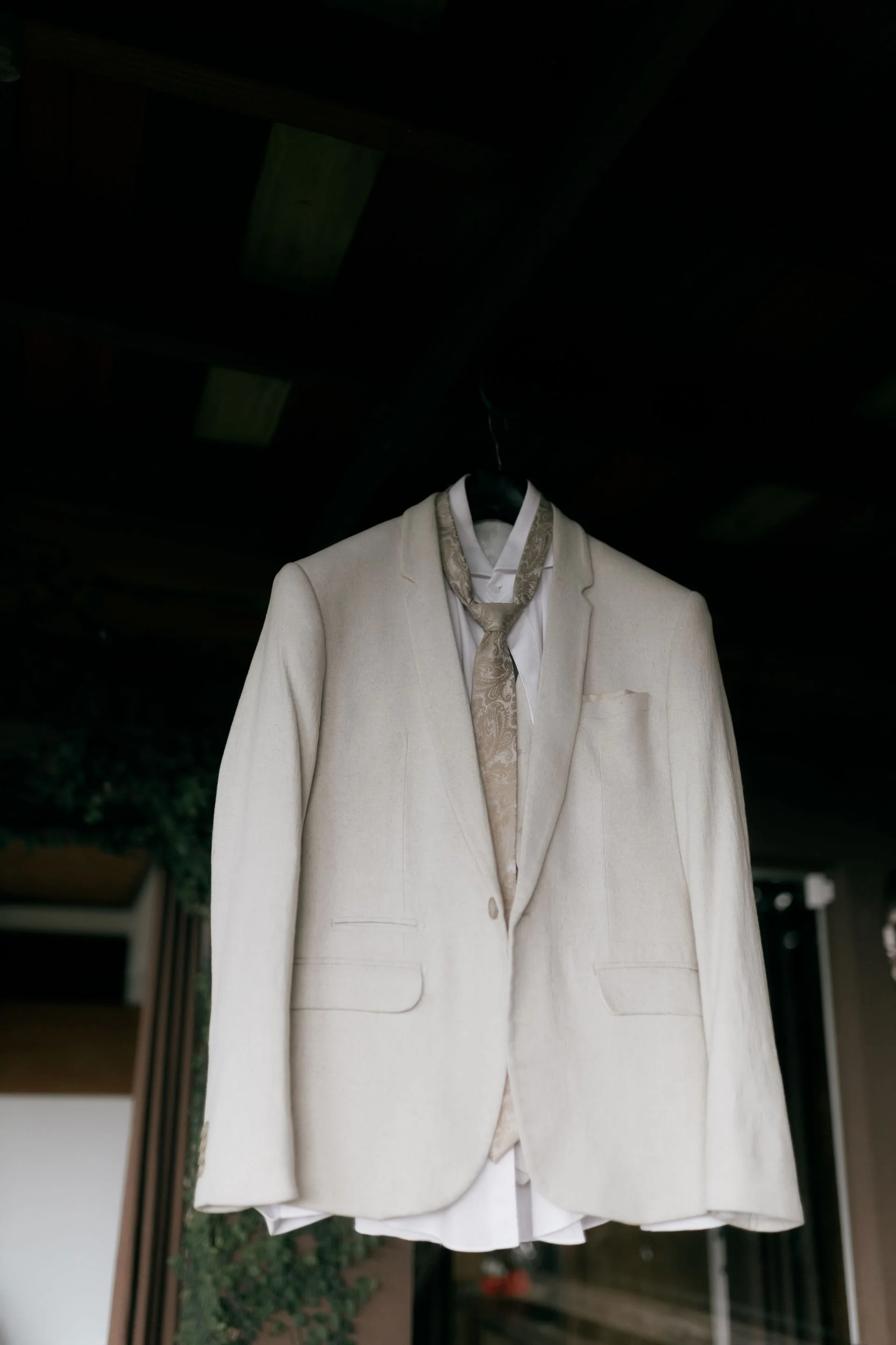 A white suit jacket with a patterned beige scarf hanging on a black hanger in front of a dark background.