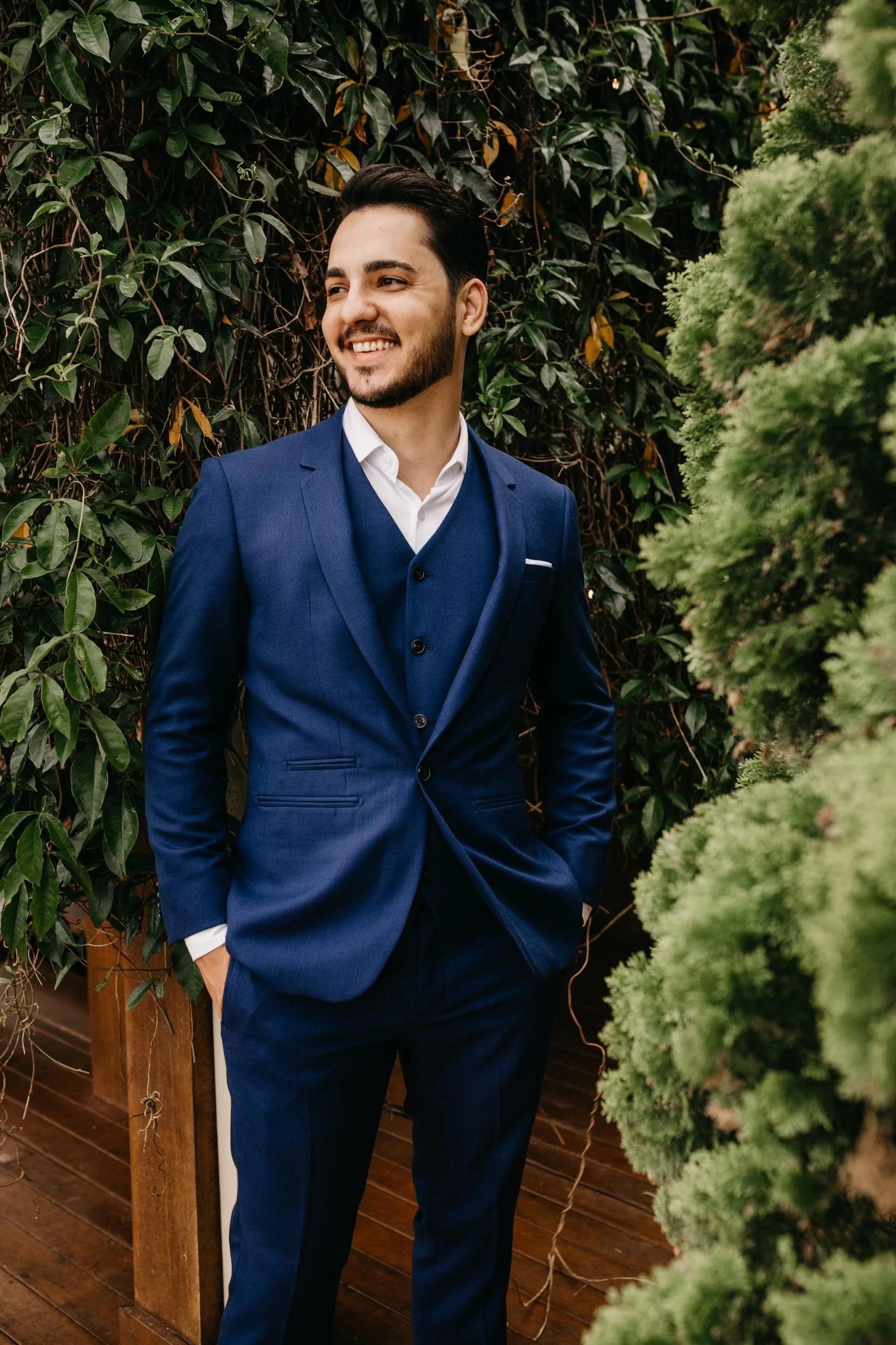 A young man in a navy suit with a white shirt smiling and looking to his left while standing outdoors among green foliage and plants.
