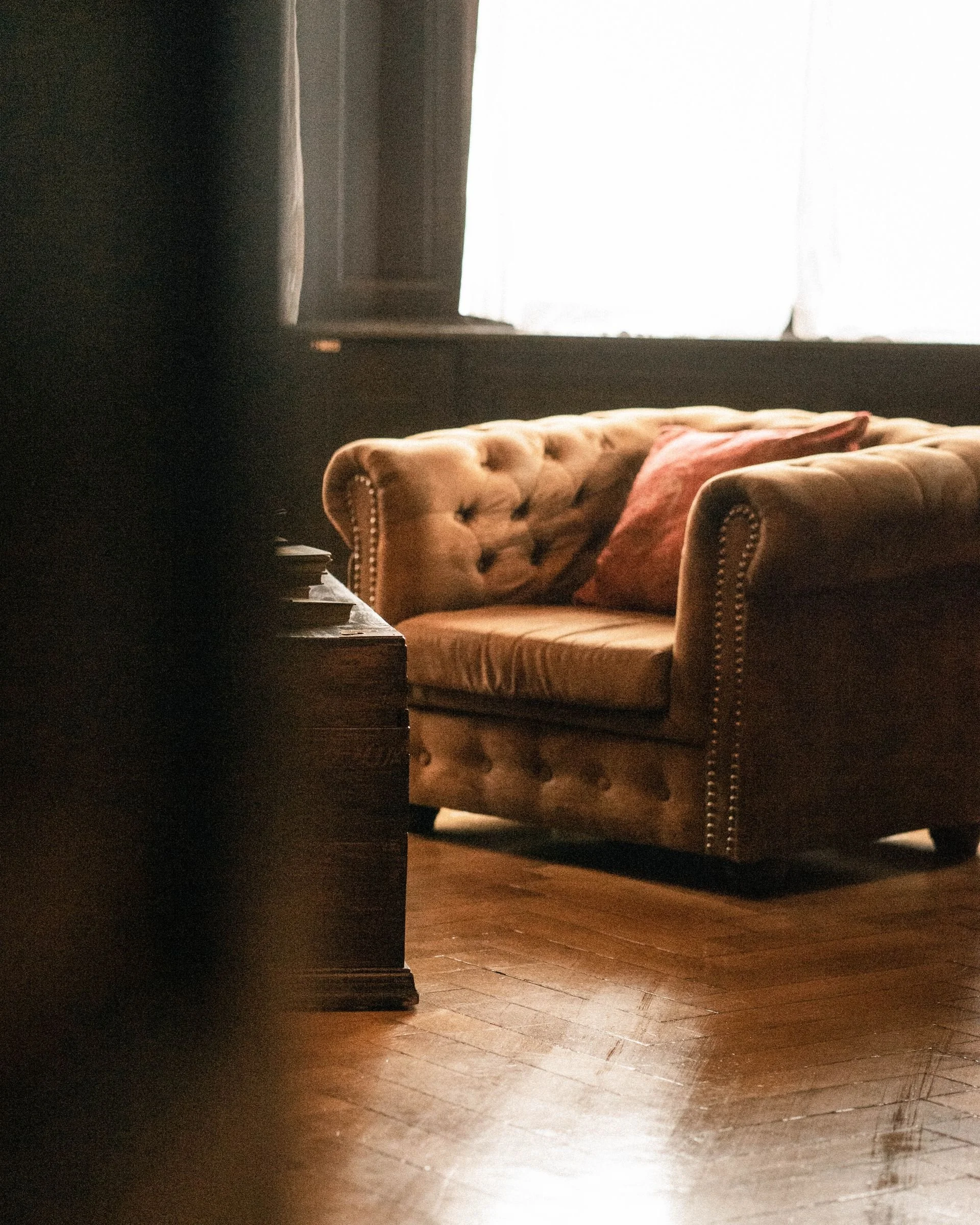 Living room with wooden floor, antique tufted sofa with a pink cushion, next to a wooden dining table, near a window with sheer curtains letting in soft natural light.