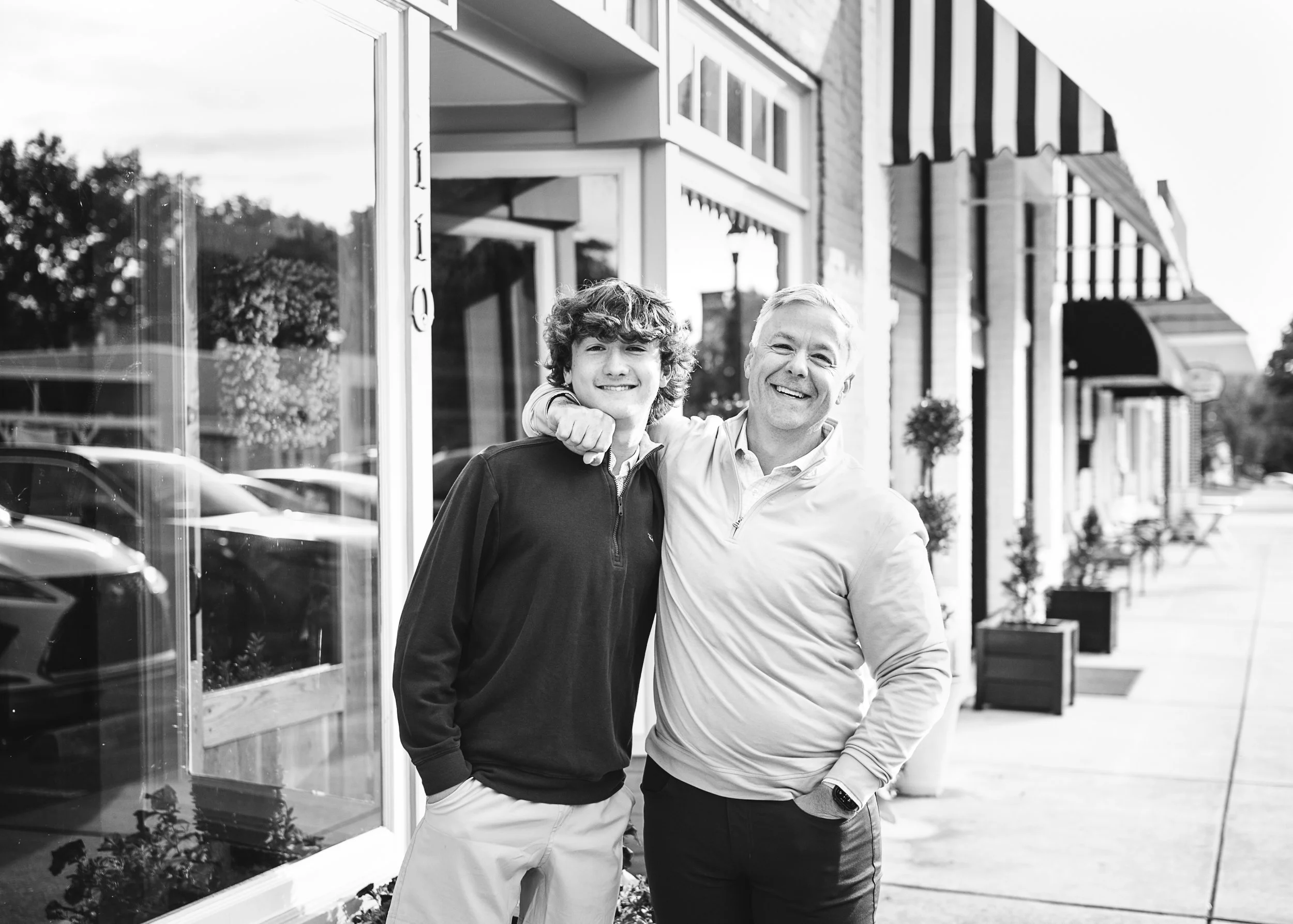 A smiling young man and an older man with a smile, standing close together outside a storefront with large windows and striped awnings, in black and white.