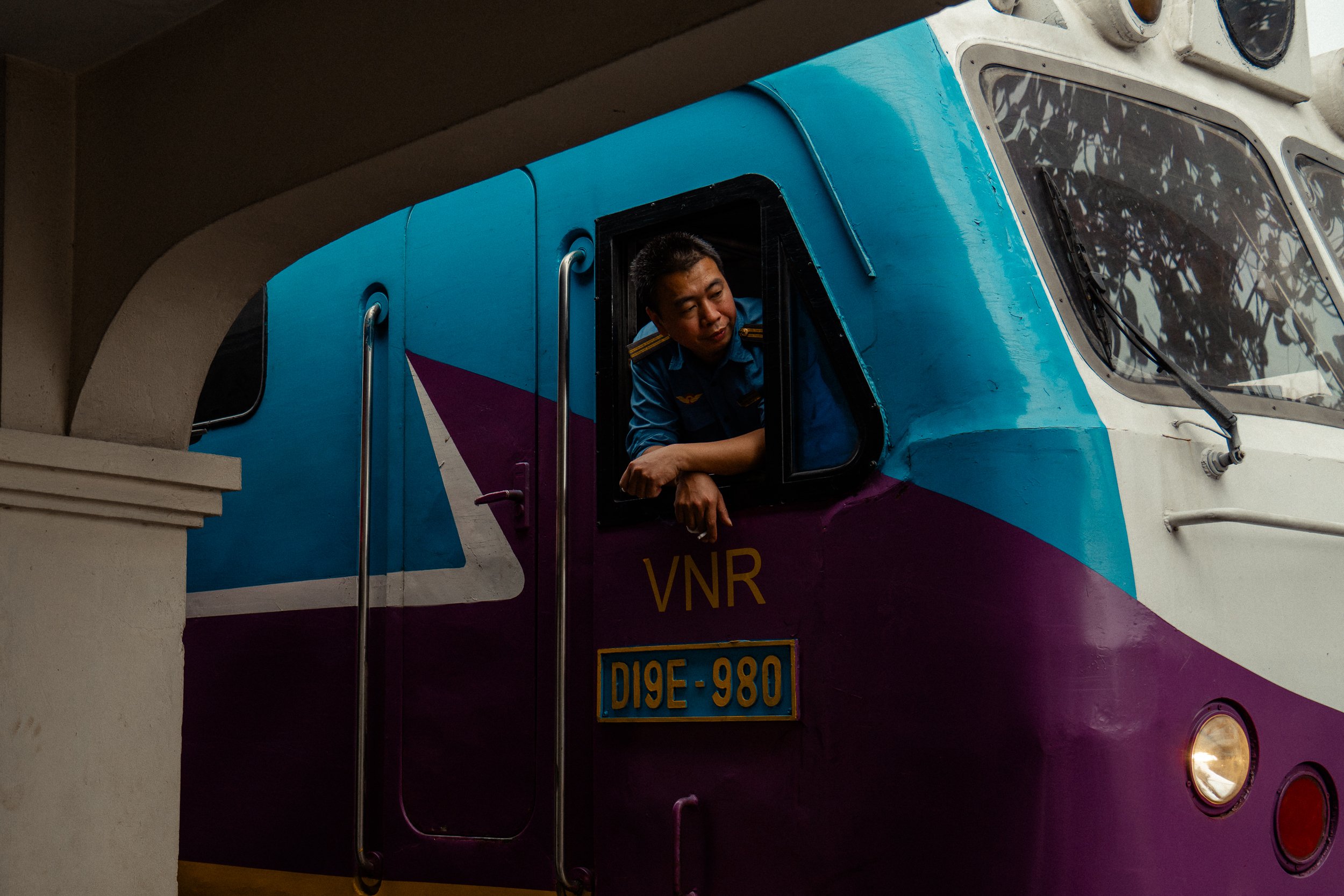 A person leaning out of the window of a colorful bus with a blue, purple, and white paint job, featuring a license plate reading D19E-980.
