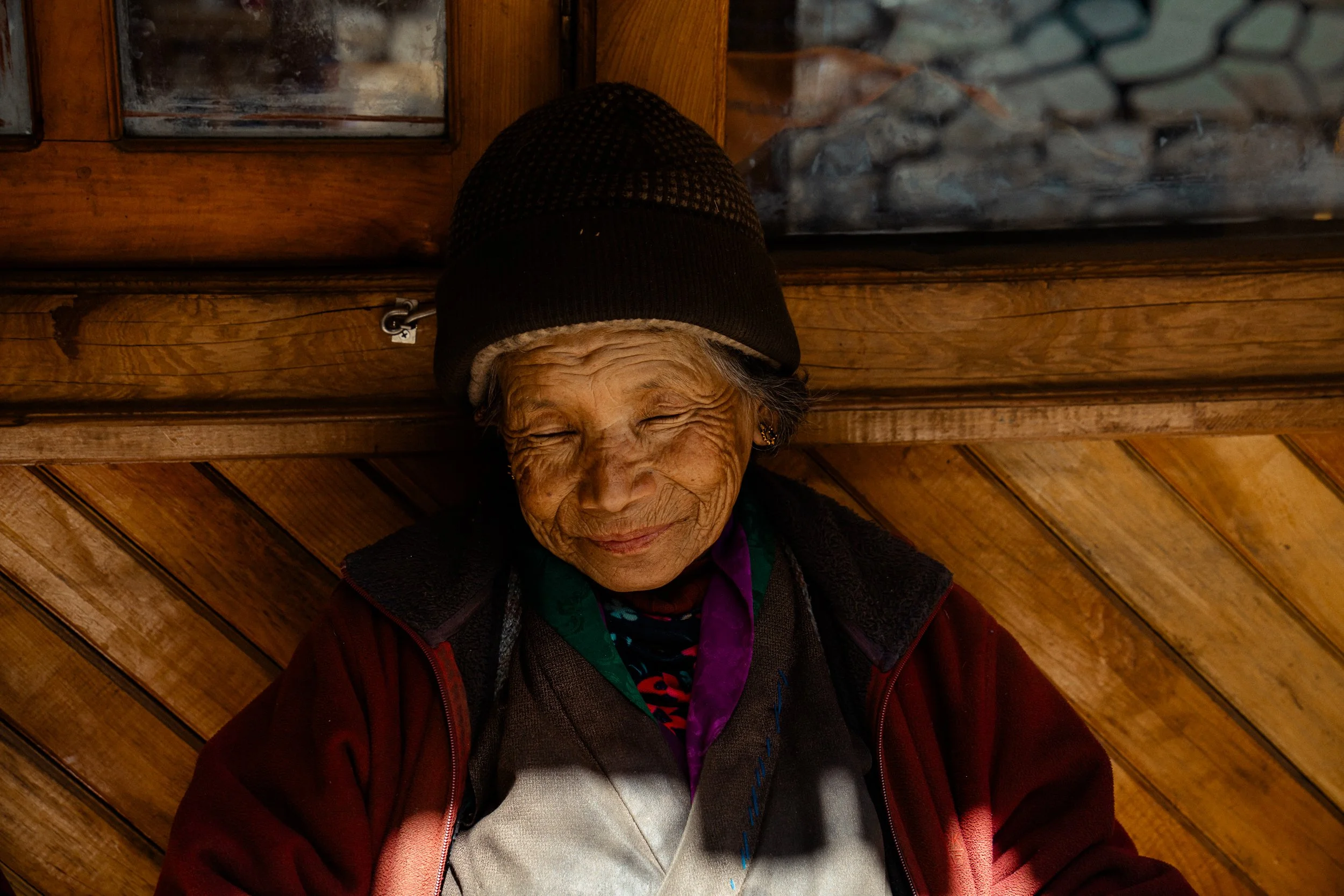 An elderly woman with gray hair and wrinkles, wearing a black and brown beanie, a maroon jacket, and a purple scarf, sitting against a wooden wall with diagonal paneling.