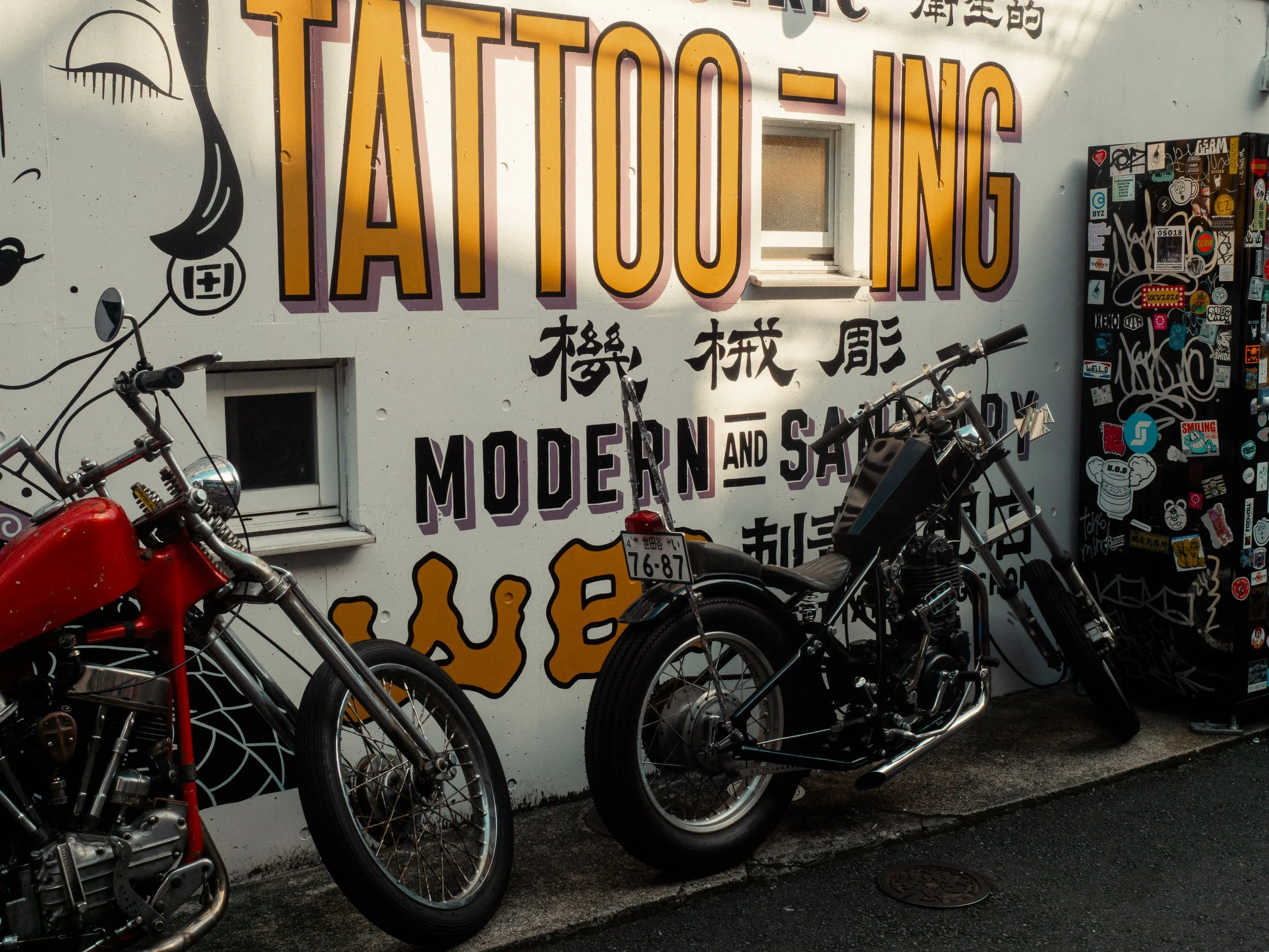 Street scene with motorcycles parked in front of a wall featuring large graffiti and stickers. The graffiti includes bold yellow and black letters and some Japanese characters. The wall also has a small window.