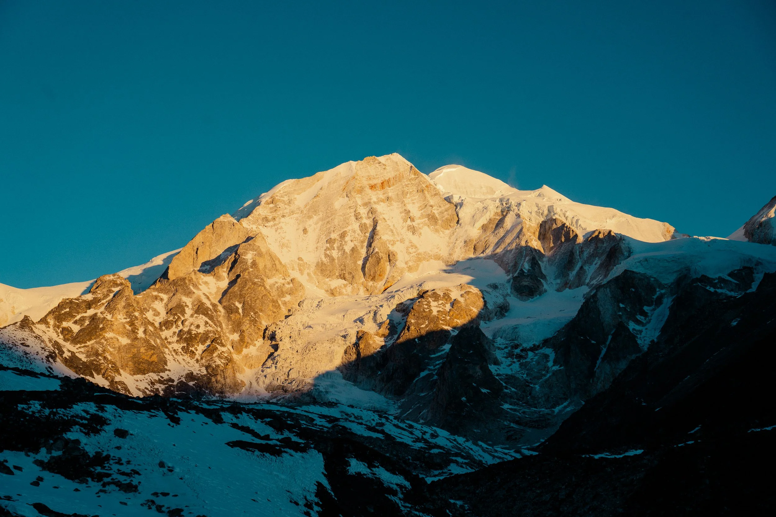 Snow-capped mountain peaks illuminated by sunlight against a clear blue sky.