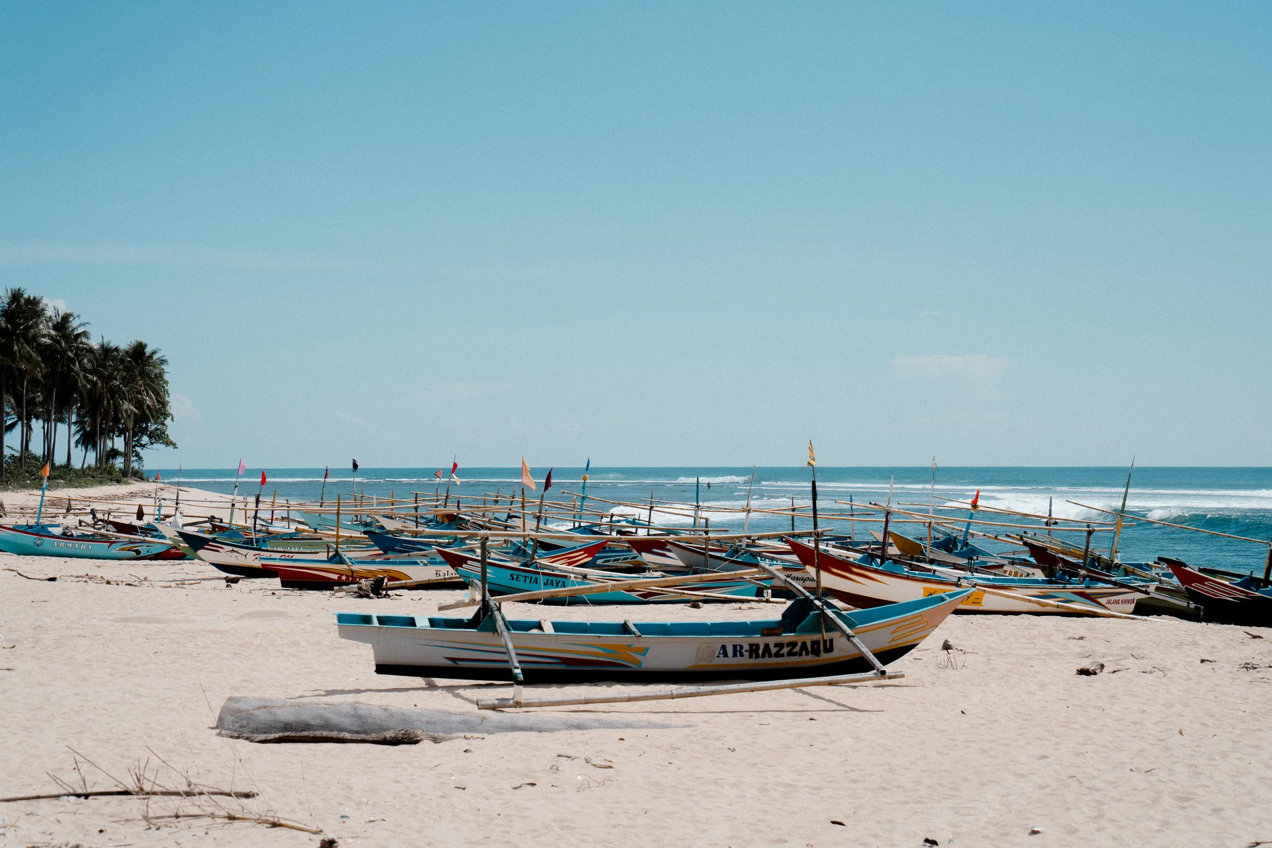 Beached traditional boats with colorful flags on a sandy beach, with a line of palm trees and the ocean in the background under a clear blue sky.
