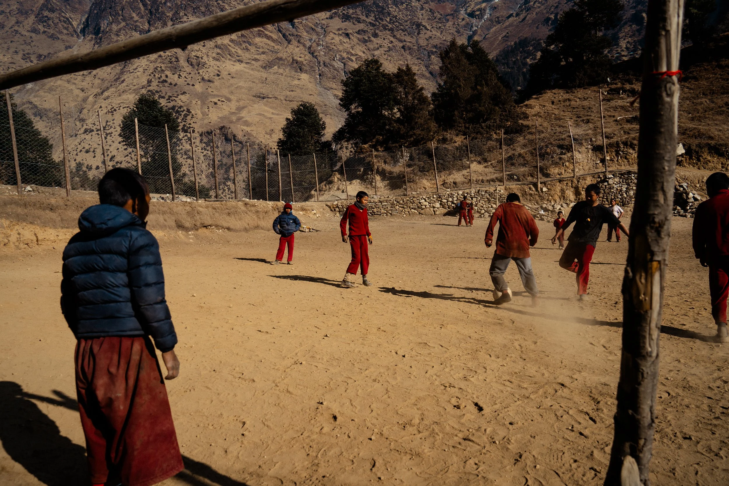 Children playing soccer on a dusty field with mountains in the background and a chain-link fence.