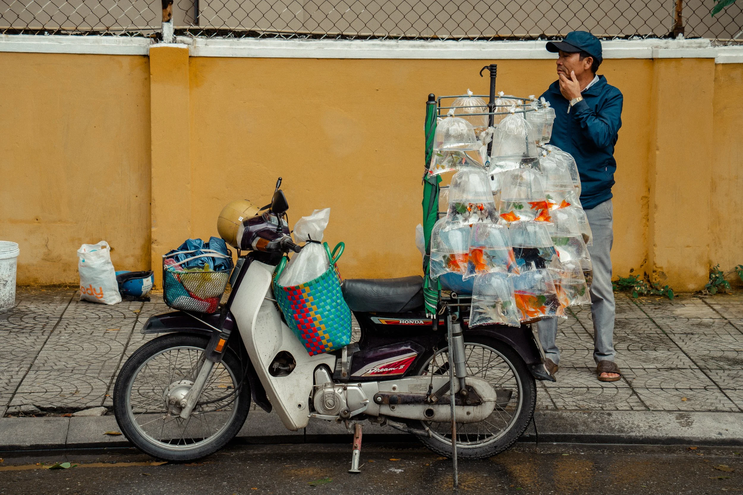 A street scene with a man standing next to a motorcycle that has a cart attached, filled with plastic bags of fish and other items. The man is wearing a dark cap and jacket, with a yellow watch, and he appears to be looking away. There is a yellow wa