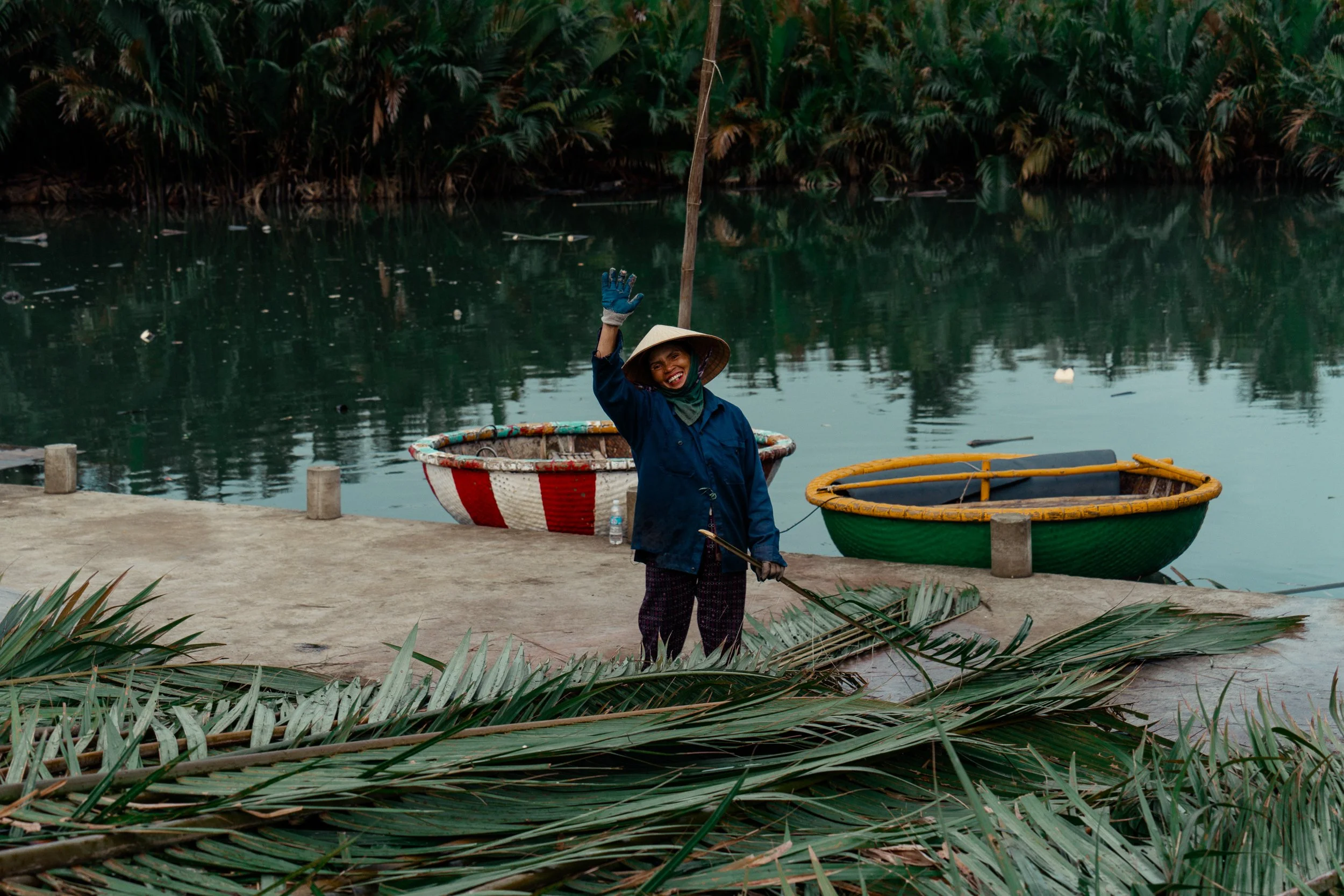 A smiling woman wearing a traditional Vietnamese conical hat, blue jacket, and gloves, waving her hand near a body of water with boats and lush green foliage in the background.