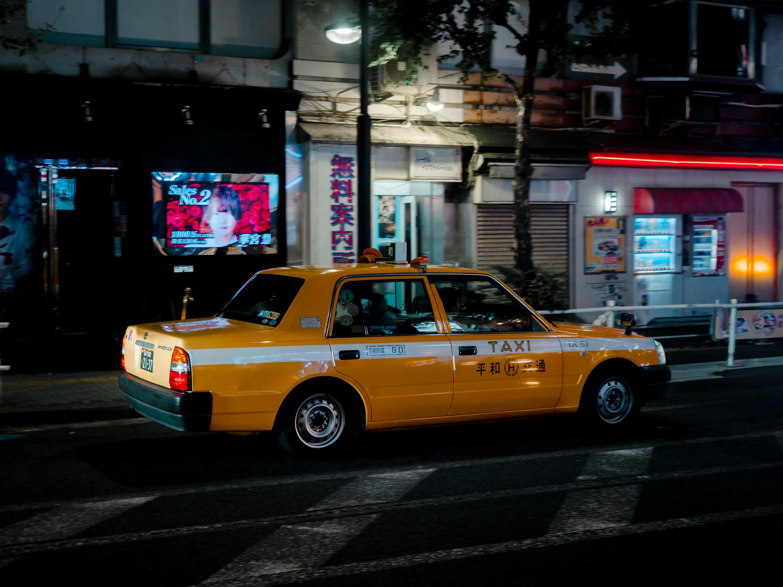 A yellow taxi cab driving at night on a city street with illuminated signs and storefronts.
