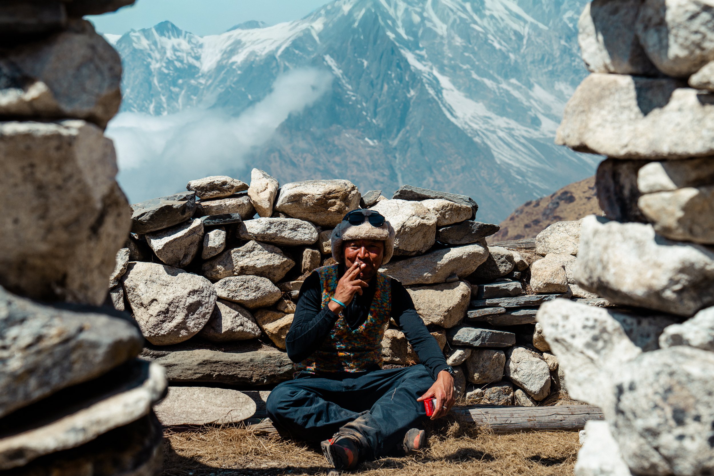 A person with a fur hat and sunglasses sitting between stone walls with snow-capped mountains in the background.