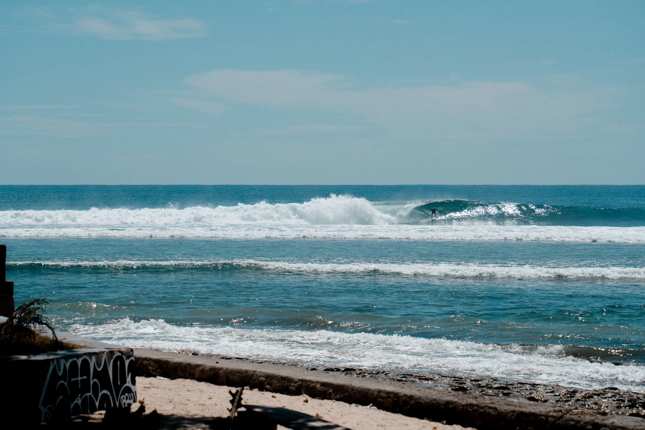 View of a beach with waves, a surfer in the water, and a graffiti-covered wall in the foreground.