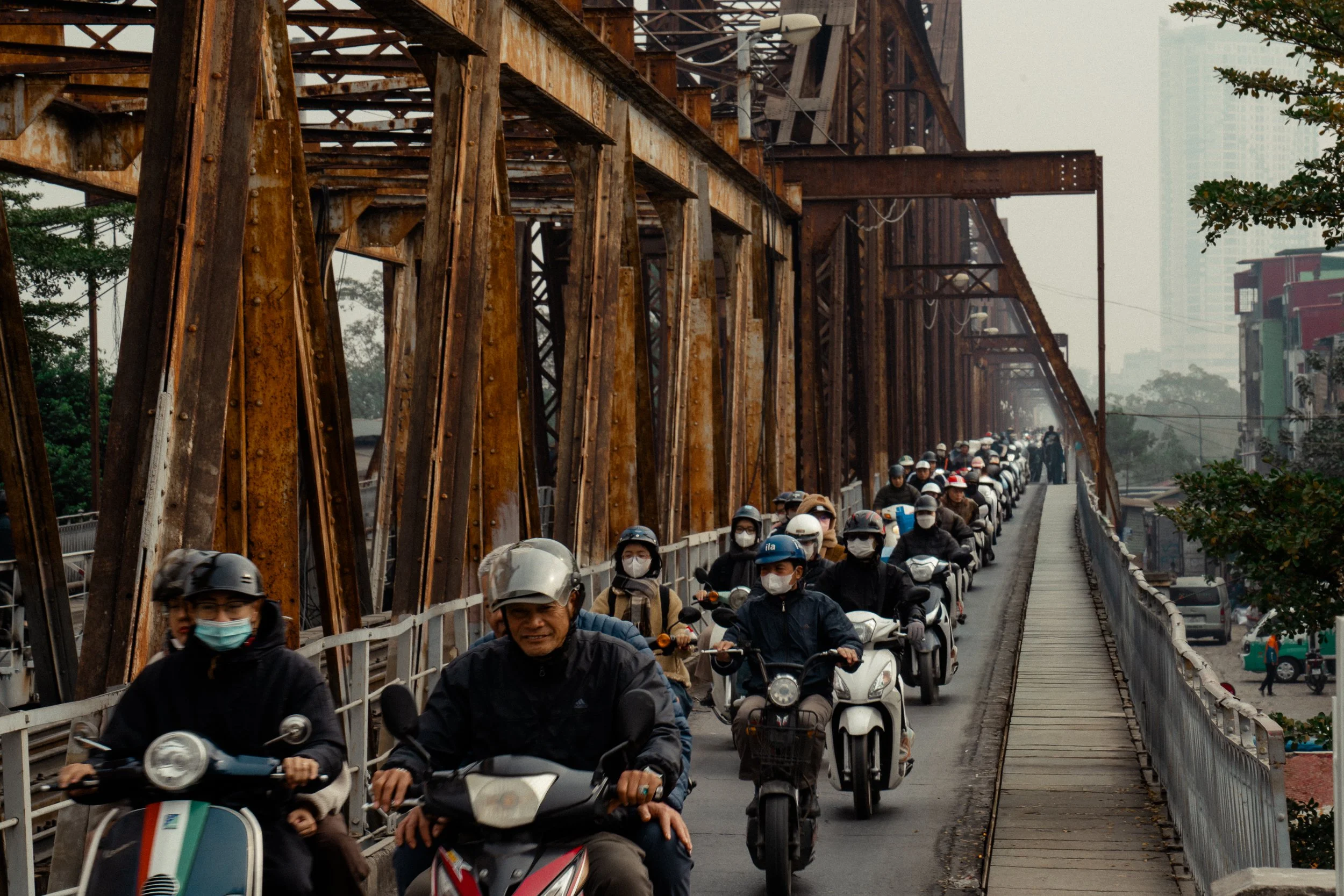 A busy bridge filled with many people riding scooters and motorcycles, many wearing face masks, with a rusty metal structure above and city buildings in the background.