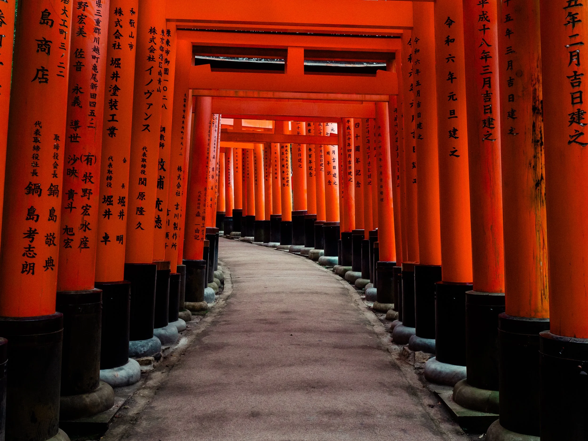 A series of red torii gates forming a tunnel at Fushimi Inari Shrine in Japan, with Japanese inscriptions on the gates and a pathway through them.