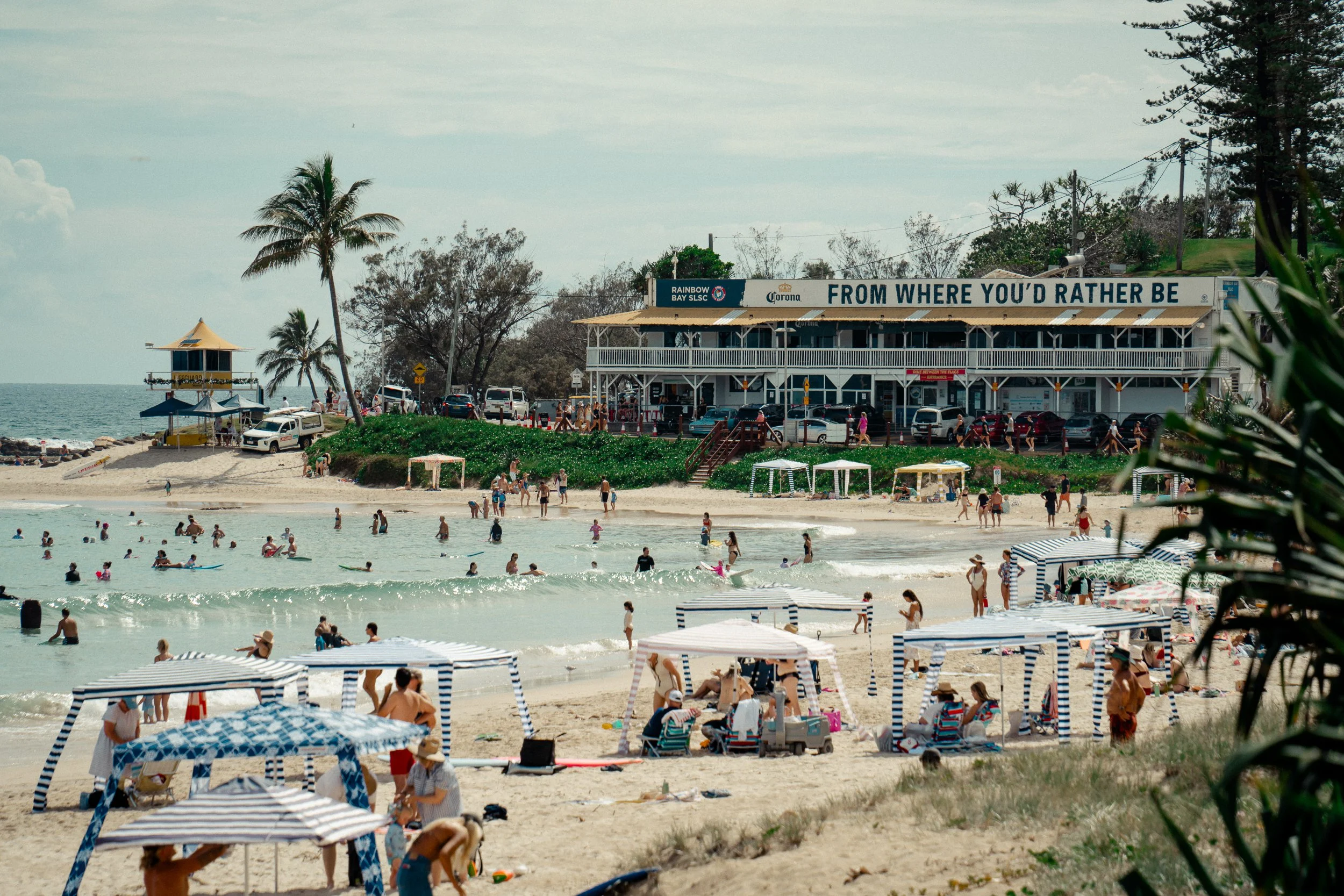 Beach with people swimming and lounging, striped umbrellas on sand, a pier with a yellow roof, and a building with a sign that reads 'FROM WHERE YOU'D RATHER BE'.